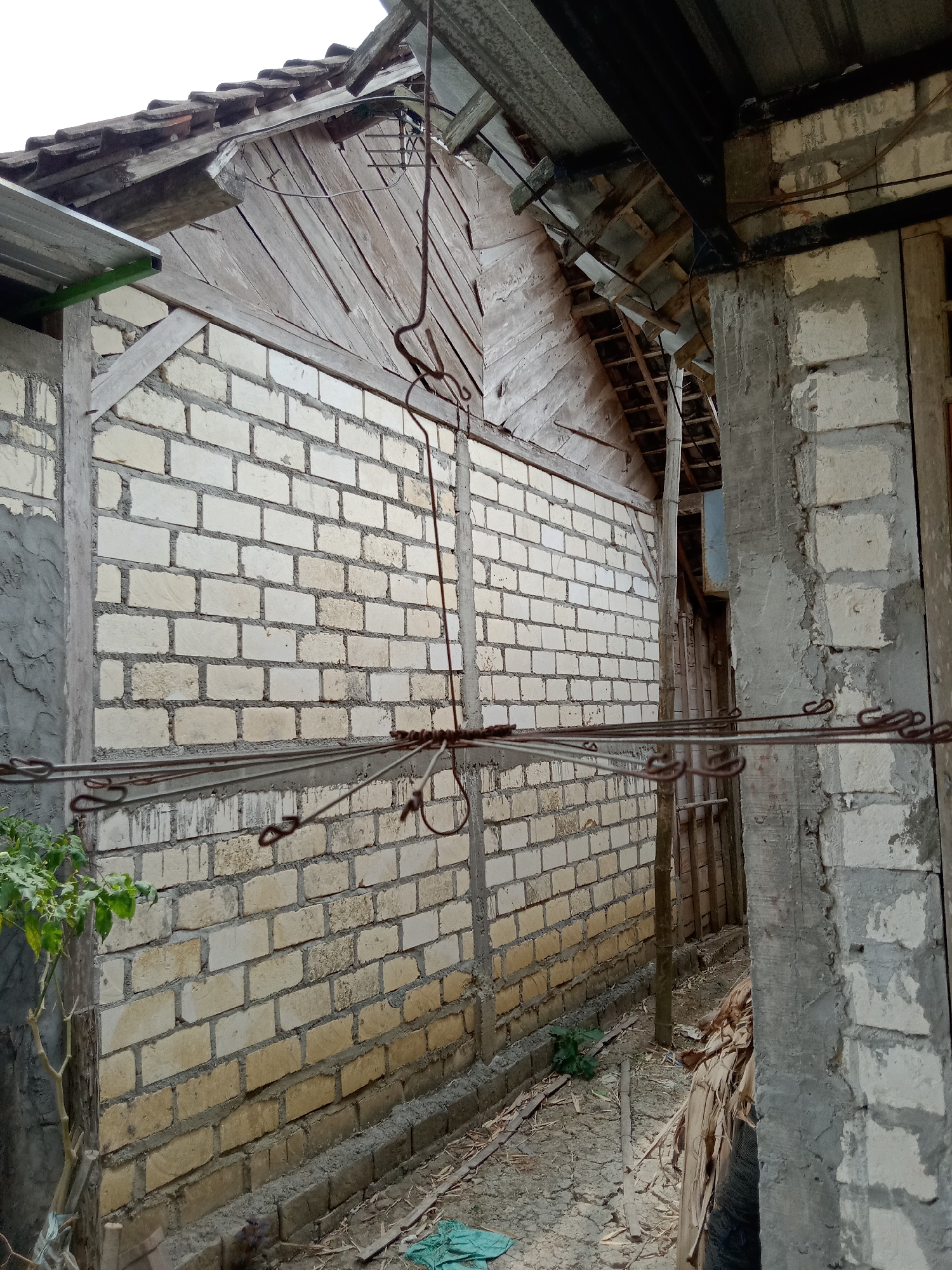The image shows a narrow alleyway between two unfinished brick buildings in Jumput, Indonesia. The walls are constructed of light-colored bricks with visible mortar. The building on the left has a wooden roof structure with corrugated metal sheeting. A metal clothesline with hooks is suspended across the alleyway. The ground is dirt and debris, with some dried leaves and a small green plant visible. The building on the right has a concrete pillar and a wooden door frame. The overall scene depicts an area under construction or in a state of disrepair.