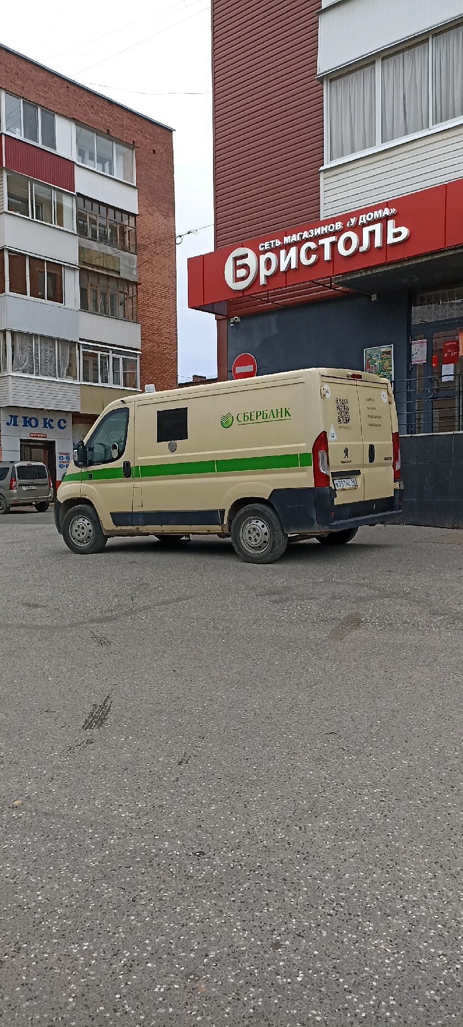 A light beige armored van with two green stripes along its side is parked on an asphalt surface in front of a building with a red facade. The van has a black bumper and wheel wells. The building has a large red sign that reads "Бристоль" (Bristol) in white lettering, with smaller text above it that says "СЕТЬ МАГАЗИНОВ. У ДОМА" (Chain stores. Near home). A window is visible above the sign. To the left of the building, a multi-story brick building with white and red elements is visible, featuring several balconies with windows. In the foreground, tire marks are visible on the asphalt. The sky is overcast. The license plate of the van is visible, reading "H757MC 182". The van is marked with "СБЕРБАНК" (Sberbank) and a logo. A QR code and other text are also present on the rear of the van. To the left of the armored van, a silver SUV is parked. A sign with the word "ЛЮКС" (Lux) is partially visible on the brick building. The general setting appears to be an urban environment in Votkinsk, Russia.