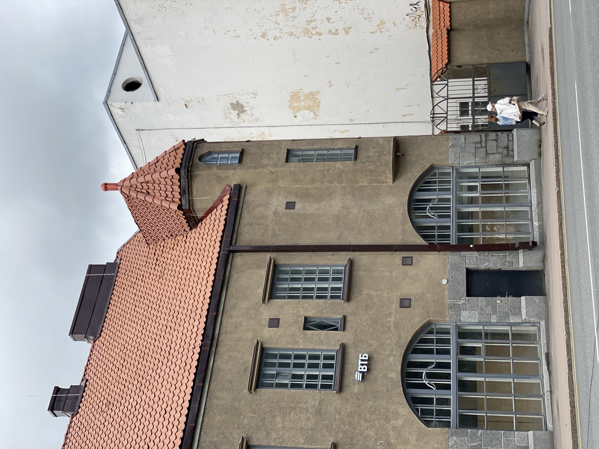 In the city of Sortavala, Russia, a brown building with a terracotta-tiled roof stands to the left, featuring large arched windows on the ground floor and rectangular windows on the upper levels. A prominent sign displaying "ВТБ" is visible on the facade. To the right, a tall, stark white building with peeling paint rises, contrasting with the older architecture. The two buildings are separated by a narrow gap. A black metal gate with decorative elements is present between the buildings, leading to an unseen courtyard. Two women are walking on the sidewalk in front of the buildings, their backs partially to the camera, one wearing a white cap and the other a blue jacket. The sky above is overcast and grey.