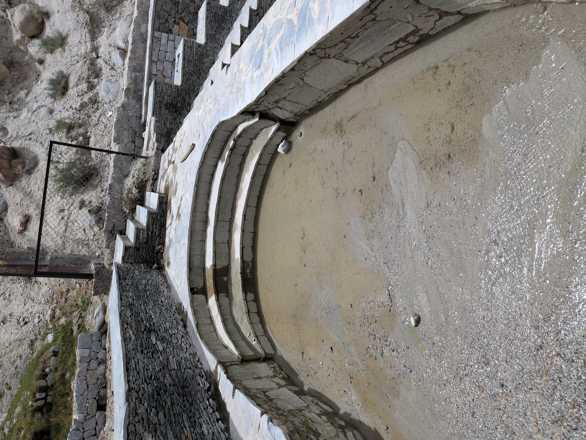 The image shows an outdoor scene in Panamik, India, featuring a partially filled, tiered pool made of stone and concrete. The pool has a curved shape with several descending steps. The water in the pool is shallow and murky, with visible sediment and small pebbles. The surrounding area is constructed with stone walls and flagstone paving. In the background, a rocky, arid hillside is visible, with sparse vegetation. A metal fence structure is also present in the background, bordering the hillside. The overall impression is of a rustic, unfinished, or natural water feature in a mountainous environment.