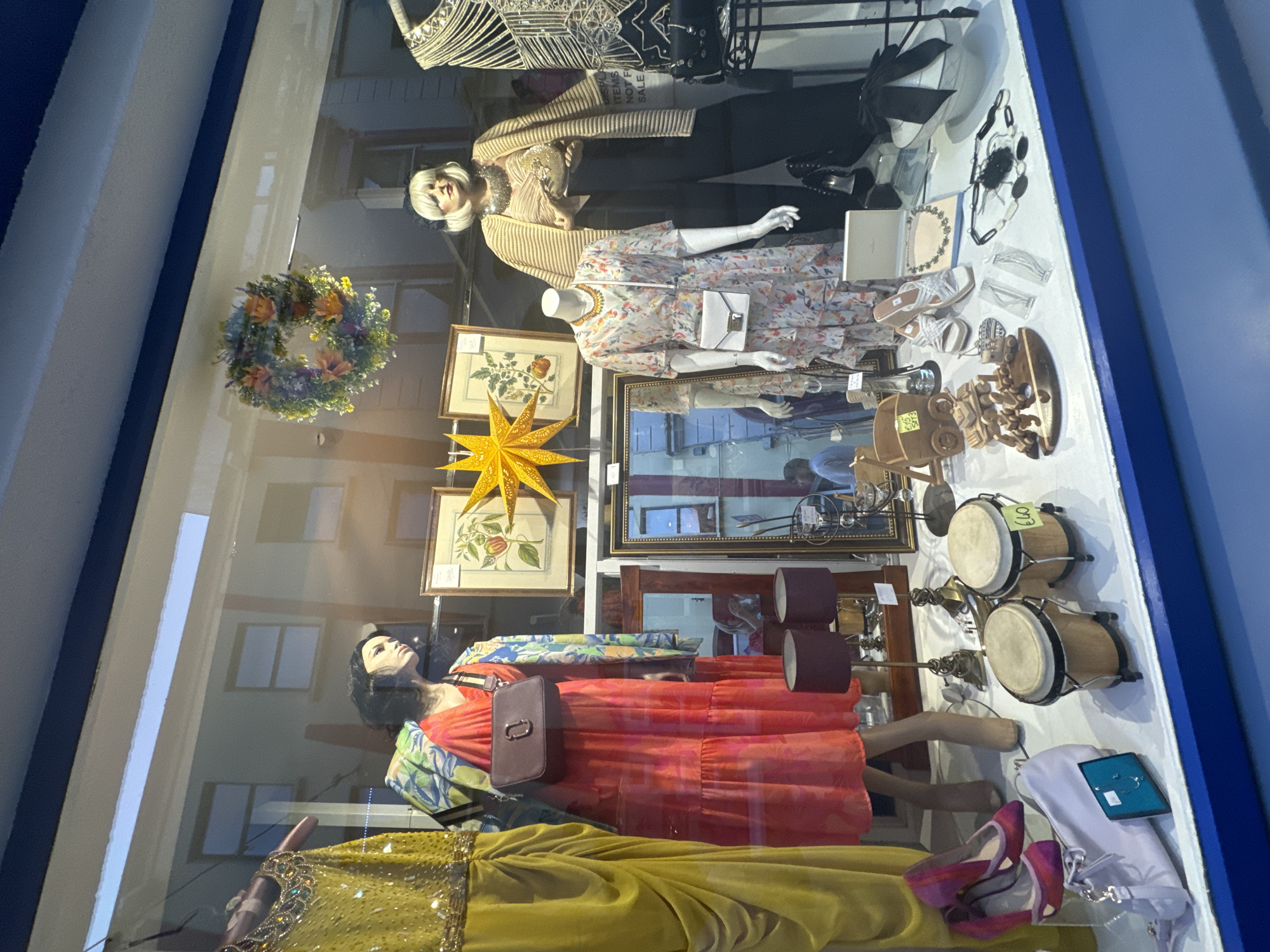 This image is a photograph of a shop window display, likely taken at a thrift store or vintage clothing boutique. The display features mannequins dressed in clothing, along with various decorative items and accessories. The shop appears to be located in Waterford City, Ireland, as indicated by the presence of a sign in the window that reads "DISPLAY ITEMS NOT FOR SALE" with "Waterford" written above it in a smaller font.
In the foreground, a pair of pink heels and a light purple handbag are displayed. Further back, there are two bongo drums with a price tag of "€40". A collection of wooden decorative items, including a small wagon and a miniature train set, are also visible. Several pairs of shoes, including white sandals and black heels, are placed strategically. Jewelry, such as necklaces and earrings, are displayed on white cards and near a white hat with a black bow.
The main focus of the display is on the mannequins. The mannequin on the left is wearing a bright coral-pink dress with a floral pattern and a light blue and green patterned jacket. It has a burgundy cross-body bag. The mannequin on the right is wearing a beige striped top, a beige cardigan, and black pants. It is accessorized with a chunky silver necklace and is holding a small, round, gold-colored purse.
Behind the mannequins, there are framed pictures on the wall, one of which appears to be a botanical illustration. A large, star-shaped yellow decoration hangs above the framed pictures. A floral wreath is also suspended in the window. The reflection of buildings across the street can be seen in the shop window, hinting at the urban environment. The lighting in the display is warm and inviting, suggesting an indoor retail setting.