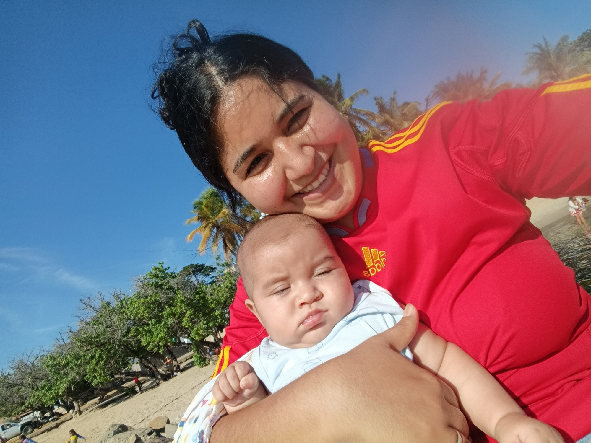 A woman is holding a baby on a beach in Juan Griego, Venezuela. The woman is smiling and looking at the camera. The baby is looking down. The background shows palm trees and the ocean. The sky is blue and clear.