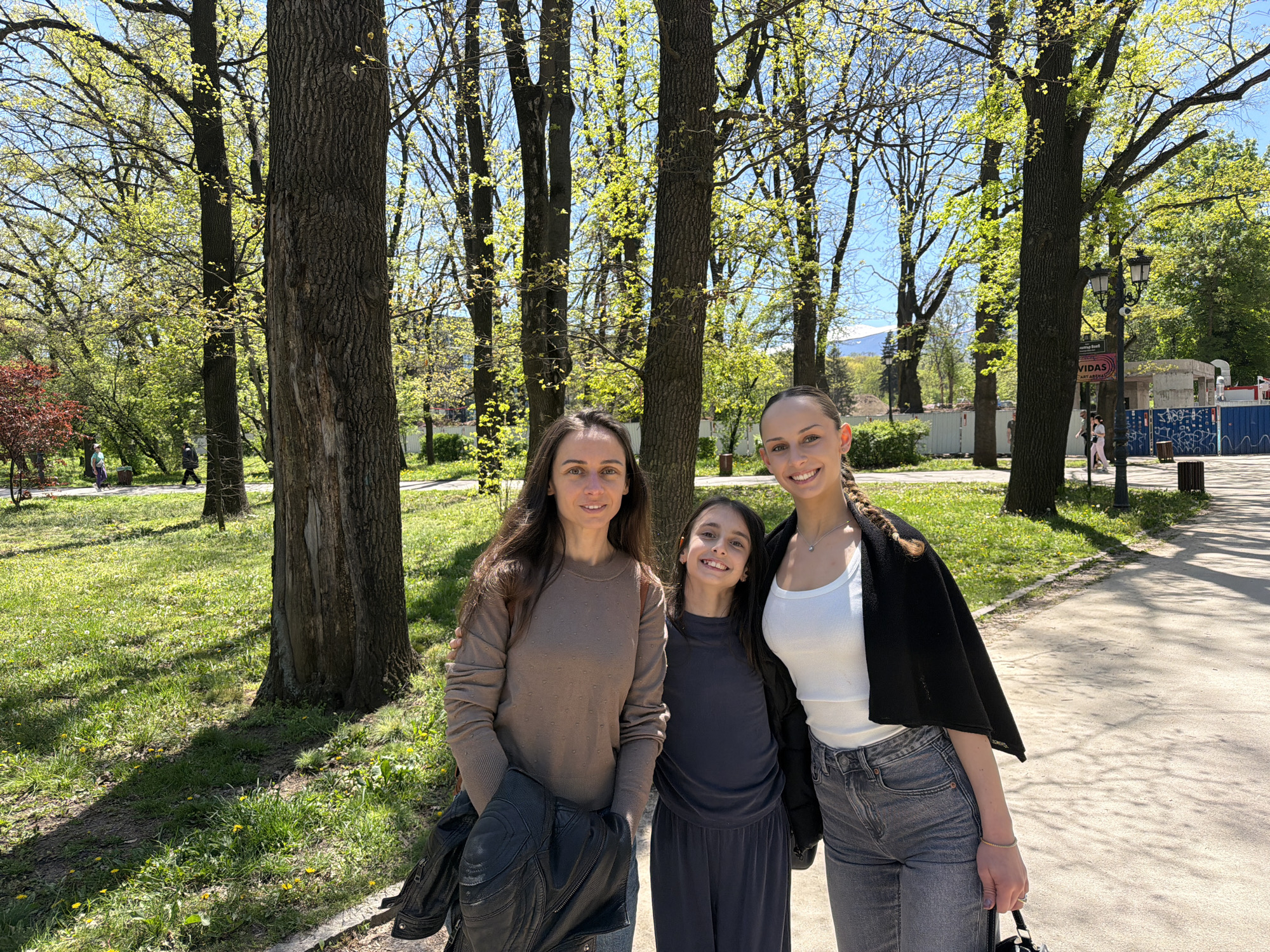 Three females are standing together in a park setting. The woman on the left has long brown hair and is wearing a tan-colored sweater with a black jacket slung over her arm. The girl in the middle is younger, with dark hair and a dark blue-grey outfit. The woman on the right has her dark hair in a braid and is wearing a white tank top, grey jeans, and a black jacket draped over her shoulders. They are all smiling. Behind them, there are numerous tall trees with green foliage. The ground is covered in grass and a light-colored pathway. In the background, there are some buildings, and what appears to be a construction area with blue panels. The general location appears to be a park within the city of Sofia, Bulgaria.