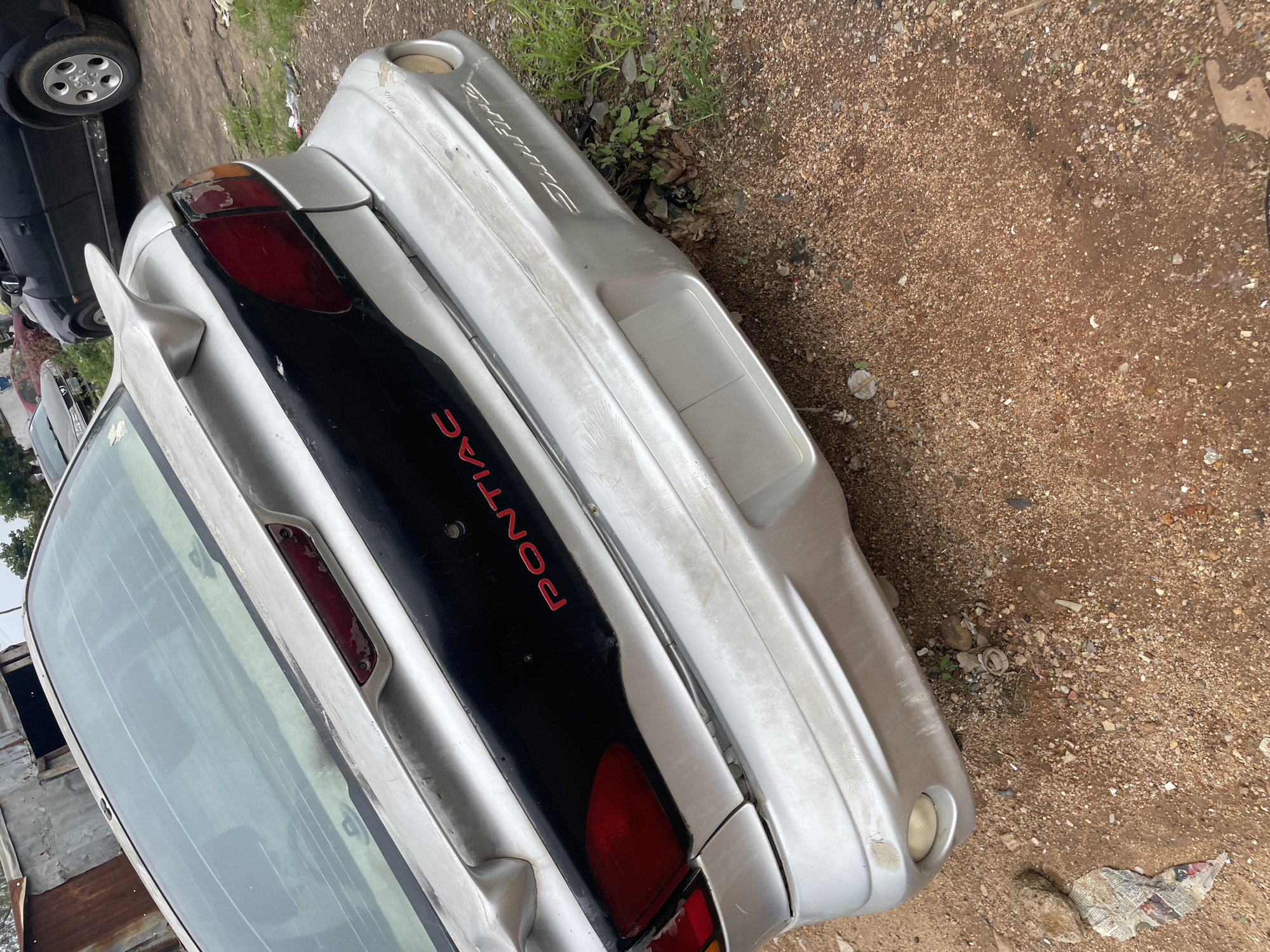 The image shows the rear view of a silver Pontiac Sunfire parked in a dirt lot. The car's paint is weathered and peeling in some areas. The car has a large black stripe across the rear, with red "PONTIAC" lettering. A spoiler is visible on the trunk. The background shows other vehicles, including a black car and a red car, and some corrugated metal structures. The ground is covered in dirt, gravel, and some sparse vegetation. The scene appears to be an outdoor storage or impound lot in New Stadium, Nigeria.