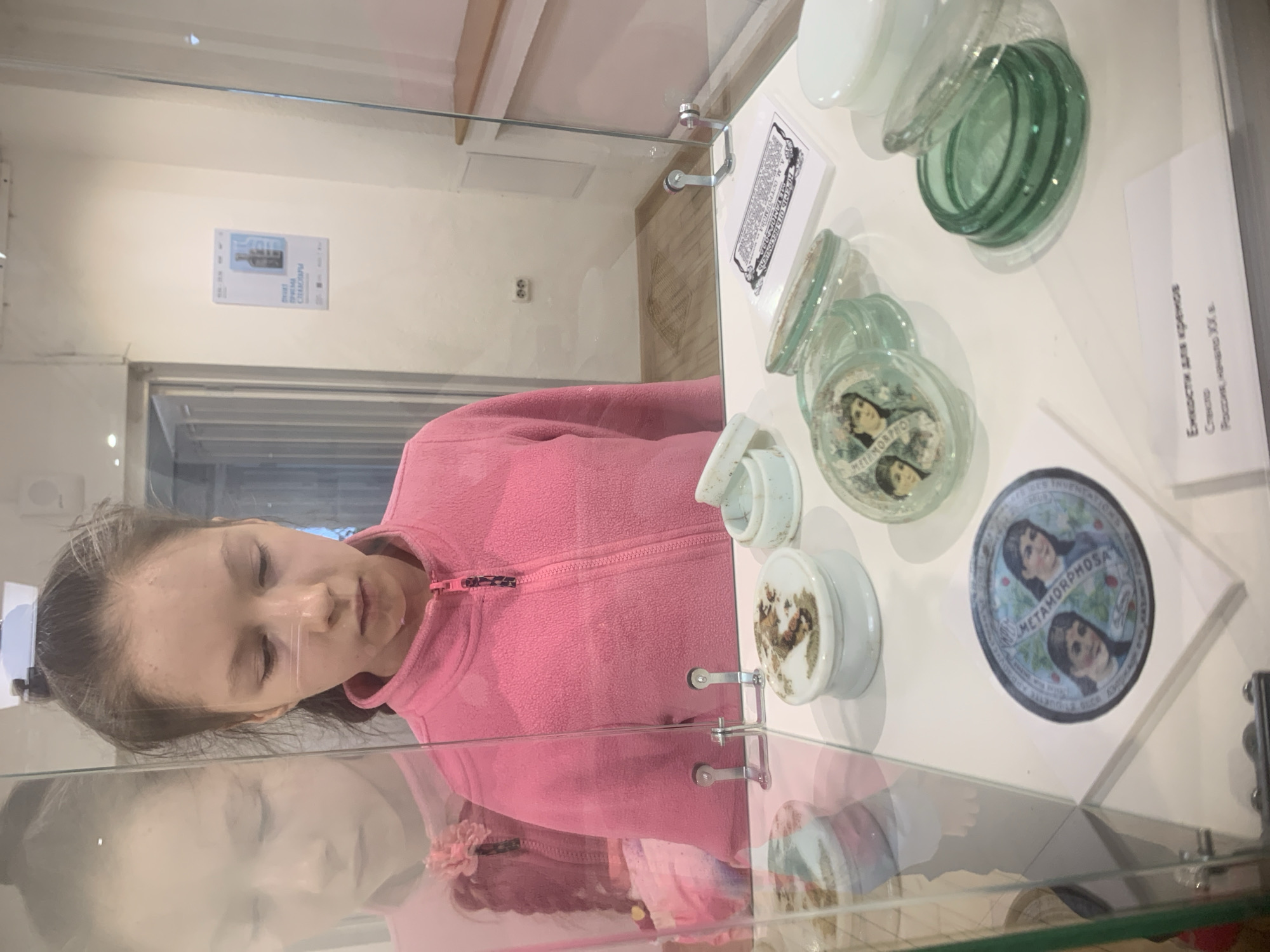 A photograph shows a young girl in a pink fleece jacket looking at a display case in a museum. The display case contains various small, circular containers, some made of clear glass, and others appear to be ceramic. One container has a picture of two women on it, labeled "METAMORPHOSA." A label next to the containers identifies them as "Емкости для кремов" (Containers for creams) made of glass in Russia at the beginning of the 20th century. There is also a poster with a picture of glass objects on the wall above the case. The reflective surface of the display case mirrors the face of the girl. There is a tag hanging from the zipper on her pink jacket. It is impossible to discern from the image if the location is in Izhevsk, Russia.