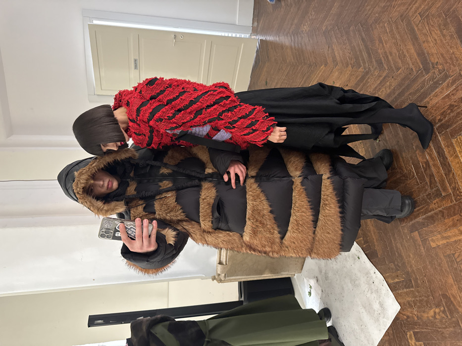 The image displays two individuals inside a bright room in Milan, Italy, characterized by dark herringbone parquet flooring and plain white walls. On the left, a male-presenting individual wears a voluminous black puffer coat with brown fur-like zig-zag panels and a fur-trimmed hood. He holds a smartphone with a patterned case, angled to capture an image of himself and the person next to him. He is dressed in dark trousers and dark shoes. To the right, a female-presenting individual, with a dark, blunt bob haircut and red lipstick, stands in profile, facing towards the smartphone. She wears a textured red and black striped jacket or top, a long draped black skirt, and black pointed-toe high-heeled boots. In the background, a white door labeled 'TOILETTE' is visible on the right. A dark clothing rack with a green garment and a crumpled white sheet are present on the far left.