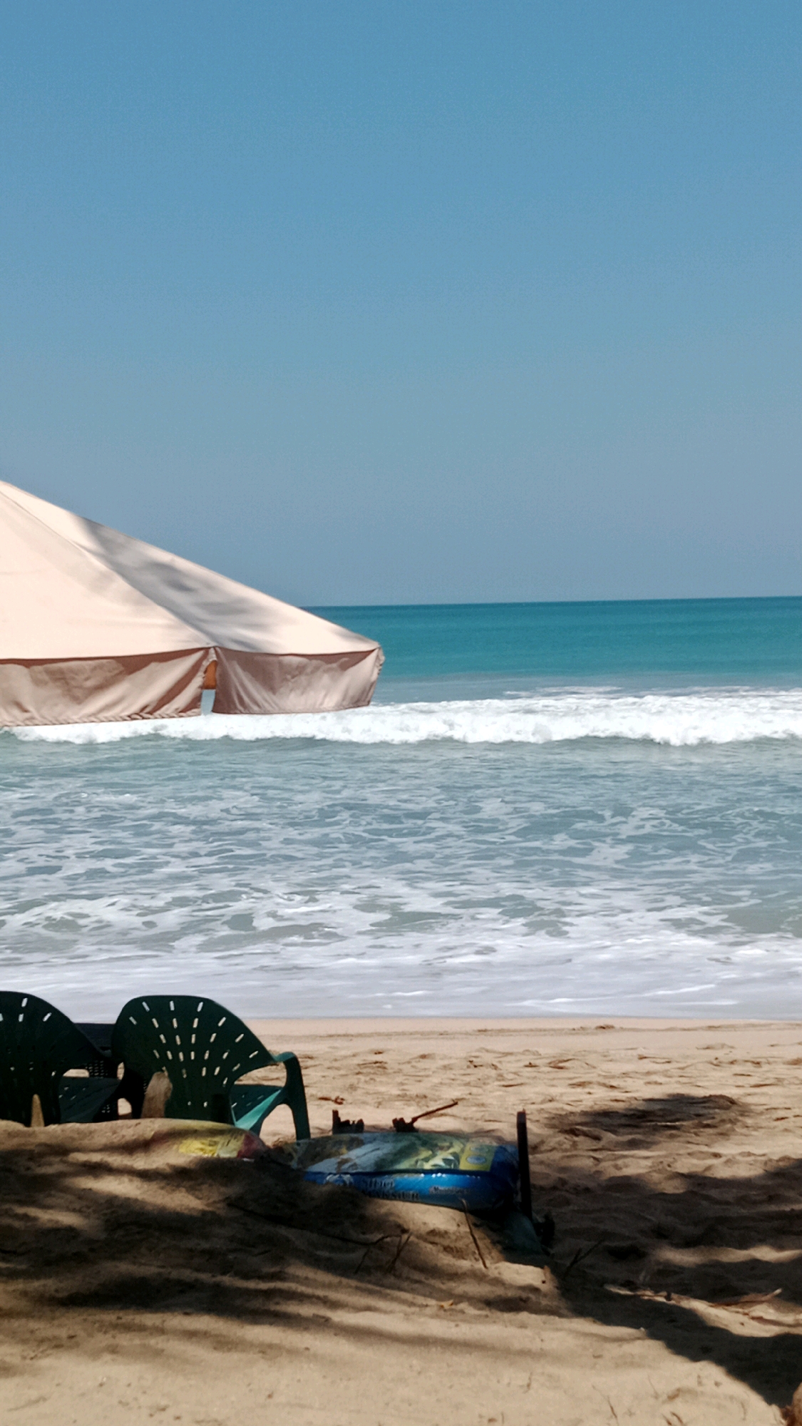 The image depicts a beach scene in Kuta, Indonesia. In the foreground, two green plastic chairs are partially obscured by sand and shadows cast by unseen foliage. A blue and yellow object, possibly a bag or mat, lies in the sand between the chairs. In the midground, gentle waves break on the sandy shore. A large, light-colored beach umbrella or tent extends from the left side of the frame, casting shadows. The ocean stretches to the horizon, meeting a clear blue sky. No people are visible in the image.