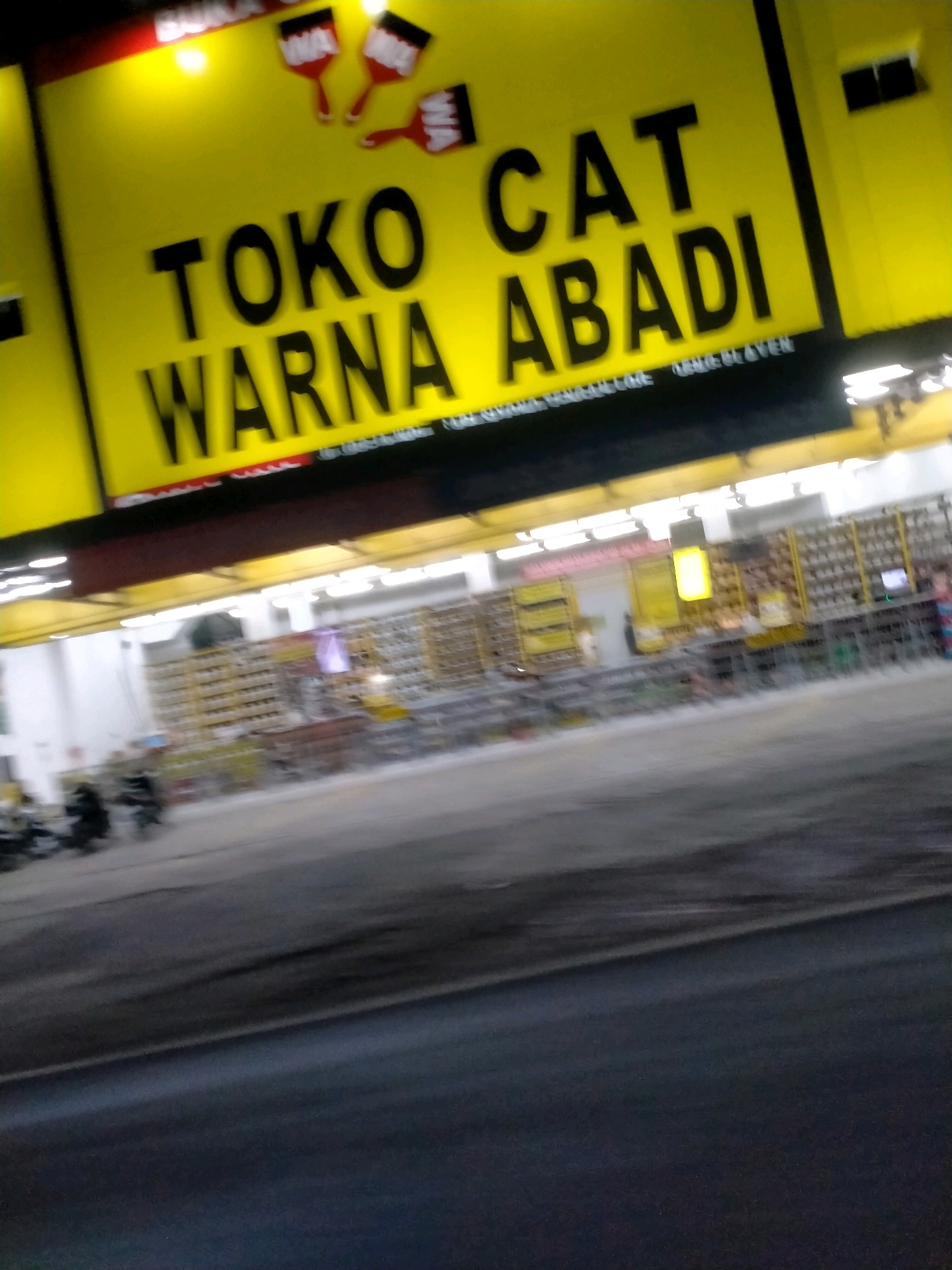 The image depicts a storefront in Playen, Indonesia, identified by a prominent yellow sign that reads "TOKO CAT WARNA ABADI" in bold black lettering. The sign also features stylized illustrations of paintbrushes. Below the sign, the interior of the store is visible, filled with shelves stacked with various products, likely paint cans and related supplies, illuminated by fluorescent lighting. A dark road is in the foreground, with a blurred depiction of a few motorcycles parked or moving to the left of the frame, indicating street activity in front of the establishment. The overall scene suggests a commercial area at dusk or night, given the artificial lighting and the dark road.