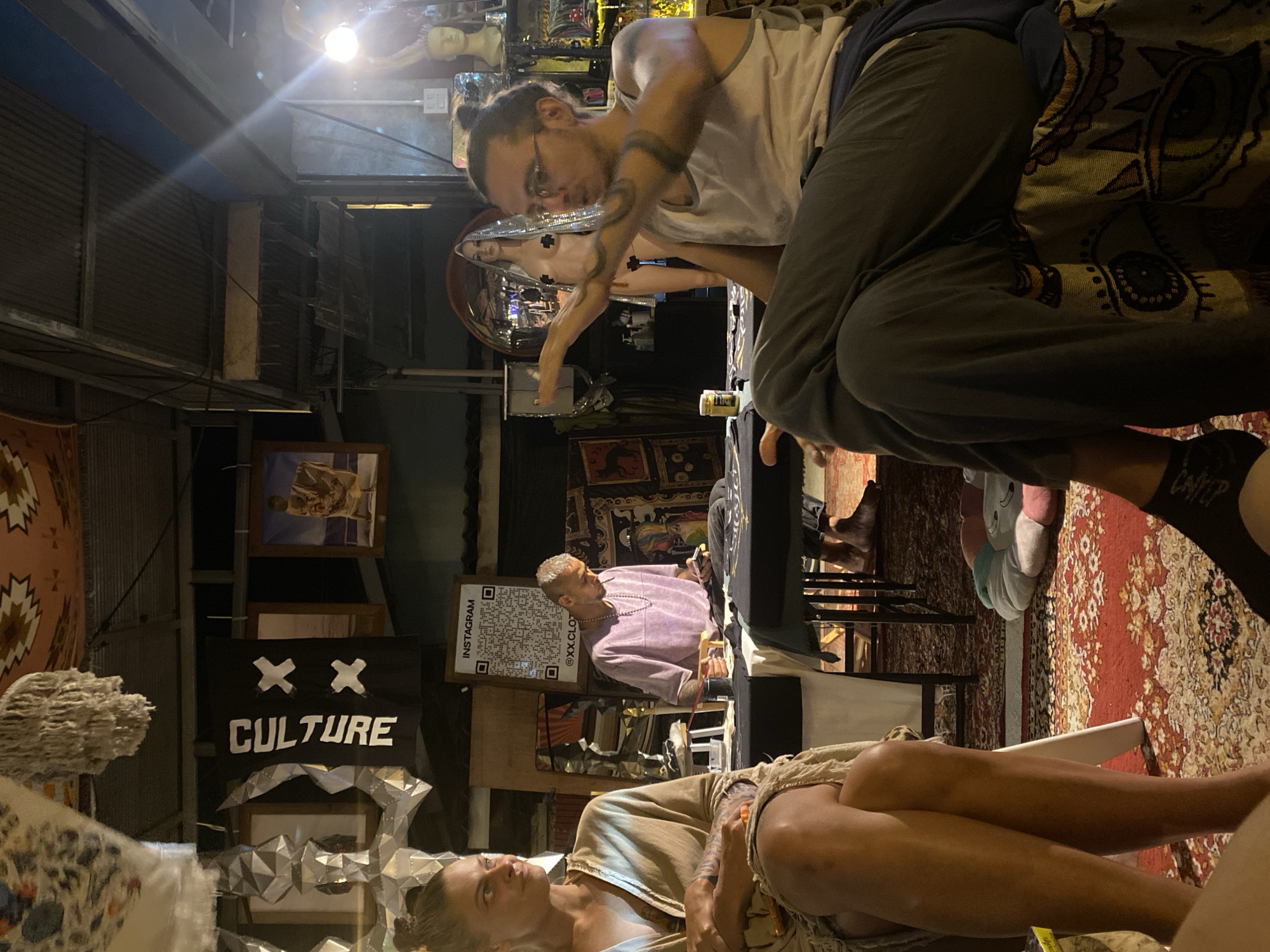 The image depicts an indoor scene with several people and objects, likely in Baan Nai Suan, Thailand.
Foreground: A person lies on a chair or surface, seemingly relaxed. Nearby, another individual sits on a higher surface.
Midground: A person with short, light hair is seated, possibly attending to a task. A banner or sign displaying "XX CULTURE" is visible. A QR code and the word "INSTAGRAM" are displayed on a rectangular sign, with what appears to be the social media handle "@XX.CLOTHING" listed below.
Background: Numerous framed photos or artworks are visible on the walls. The space appears cluttered with various decorative items.
The overall lighting is moderate, casting shadows in certain areas. The setting suggests a shop or exhibition space in Baan Nai Suan, Thailand. The environment hints at artistic or cultural activities.