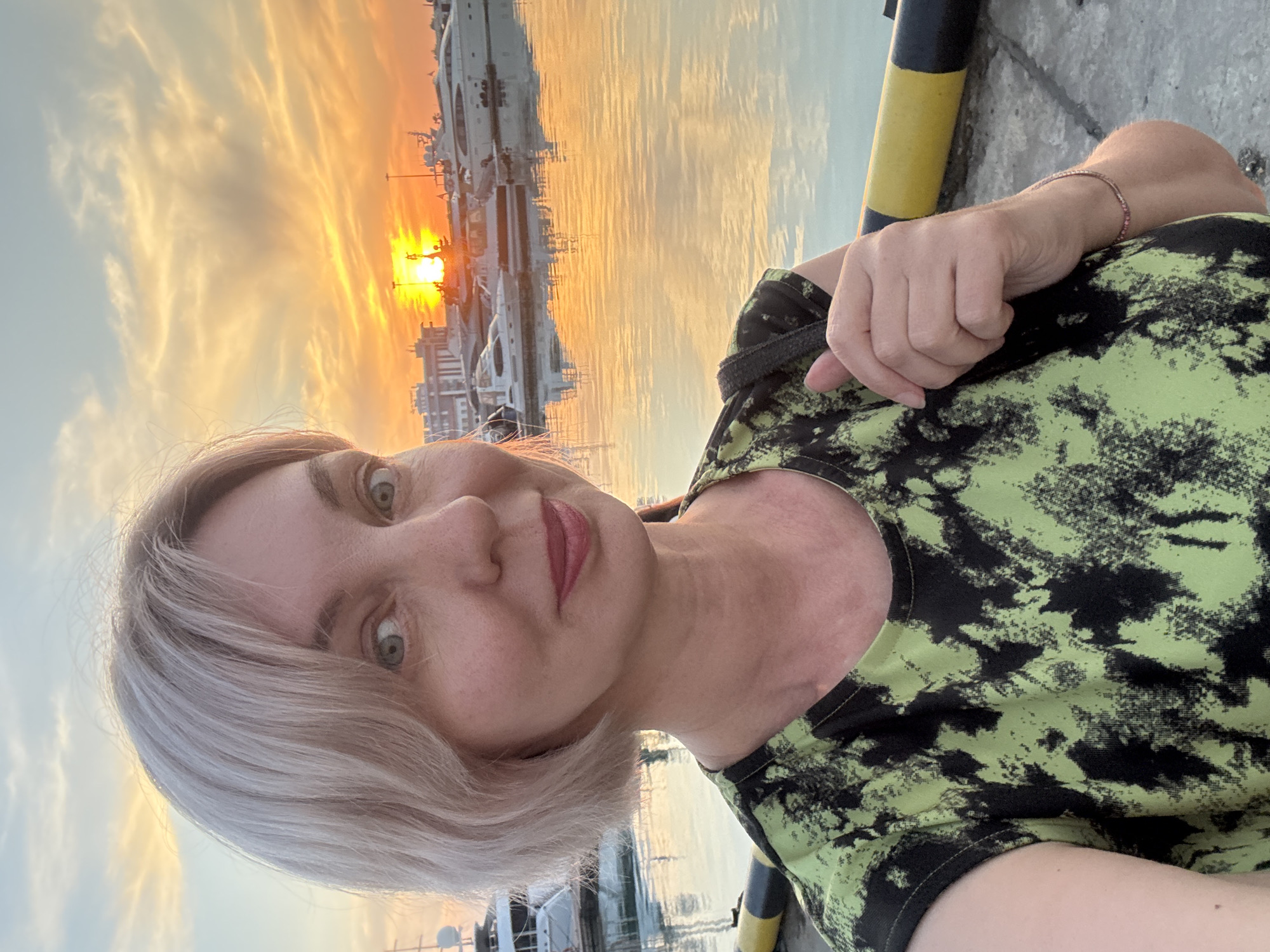 A middle-aged female presents a selfie-portrait at a marina in Sochi, Russia, during sunset. The woman, positioned centrally, has short, light blonde hair and light-colored eyes, and exhibits a slight smile while looking directly at the camera. She is wearing a short-sleeved top with a distinctive lime green and black abstract or tie-dye pattern. A thin, reddish-toned bracelet is visible on her right wrist, and her right hand grips a dark strap, possibly from a bag. The background features a calm body of water that reflects the vivid orange and yellow light of the setting sun. The sun, a bright orb, is visible near the horizon, partially obscured by distant buildings. Several large white yachts and smaller boats are moored in the marina. The immediate foreground comprises a concrete pier, featuring a black and yellow striped bollard positioned to the right of the woman. The sky overhead transitions from the intense sunset colors near the horizon to lighter blue-grey higher up, with visible cloud formations.