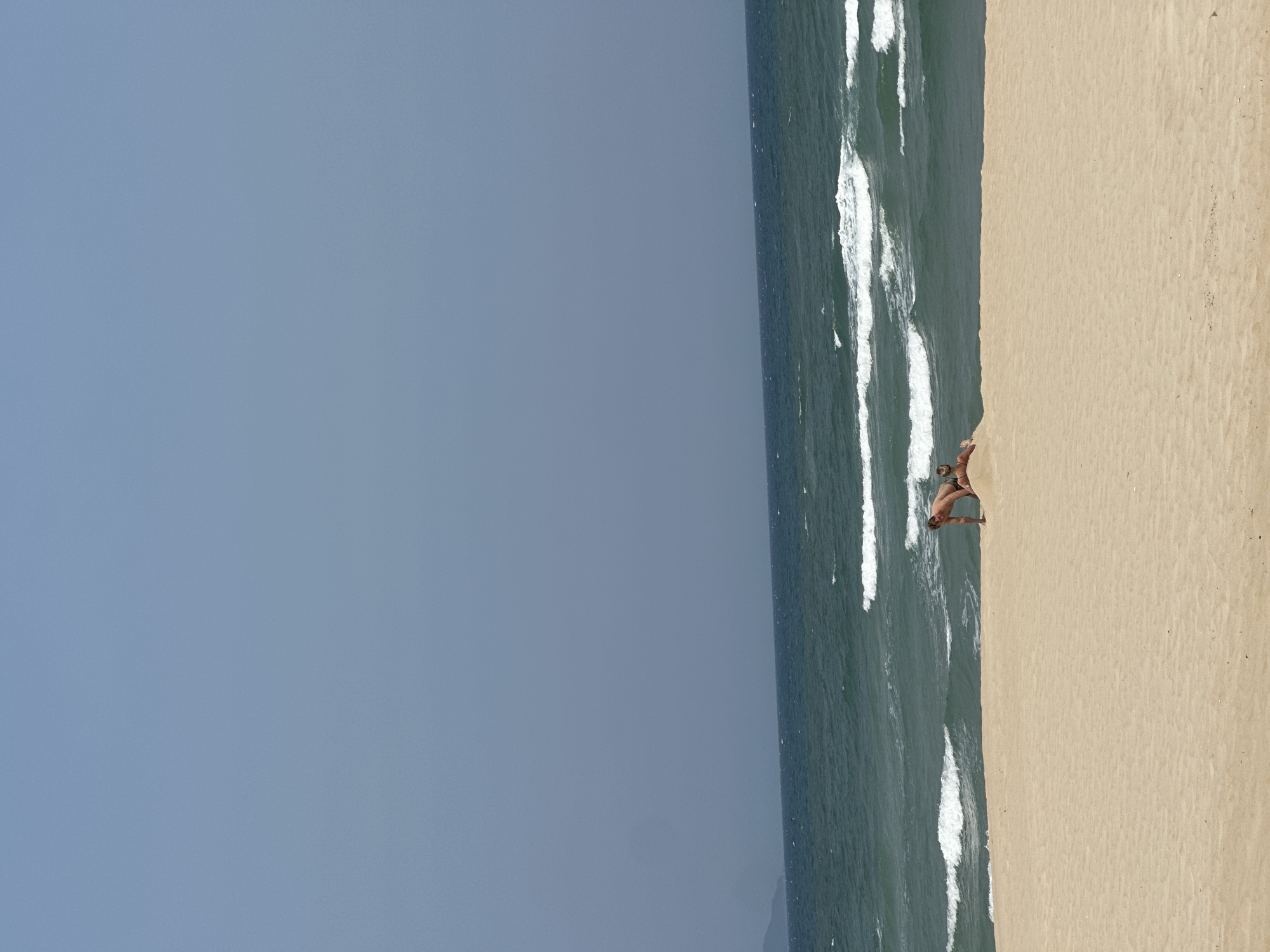The image shows a beach scene in Đà Nẵng, Vietnam. The scene is divided into thirds: the sky, the ocean, and the beach. The sky is light blue. The ocean is dark blue with white waves. The beach is tan. There is one person on the beach, a male, standing near the water's edge.