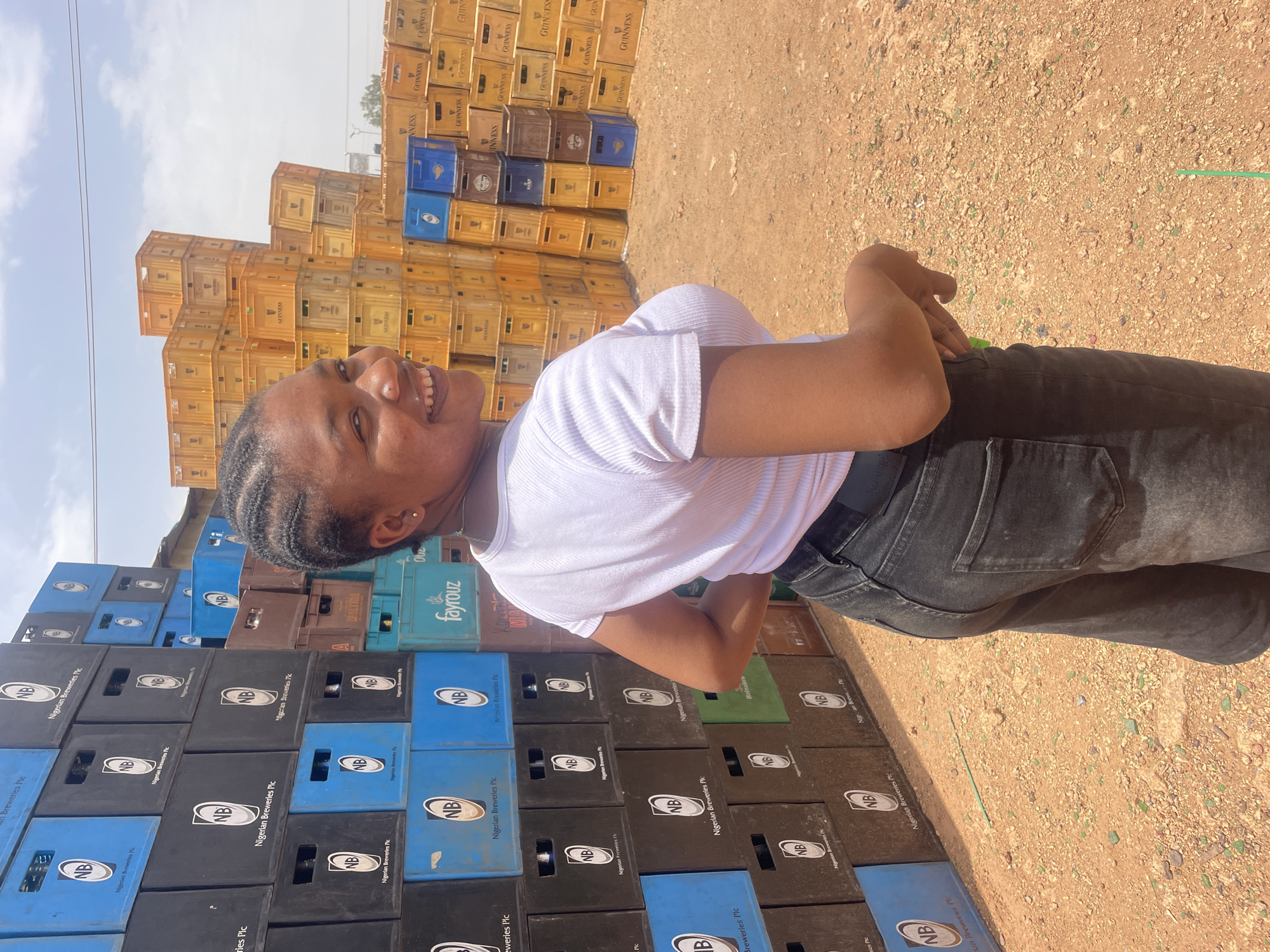 The image shows a young woman standing outdoors, positioned in front of stacks of beverage crates. The crates are stacked high, reaching several layers in height. They appear to be made of plastic and are primarily in colors of black, blue, brown, and yellow. Some of the crates feature the logo of "Nigerian Breweries Plc", while others have "Guinness" printed on them. The woman is wearing a white top, dark-colored jeans, and has her hair braided. She is smiling and looking towards the left. The ground appears to be a mix of dirt and small rocks. The sky is visible in the background, appearing bright with some cloud cover. This location is located in the city of Gura Top, Nigeria.