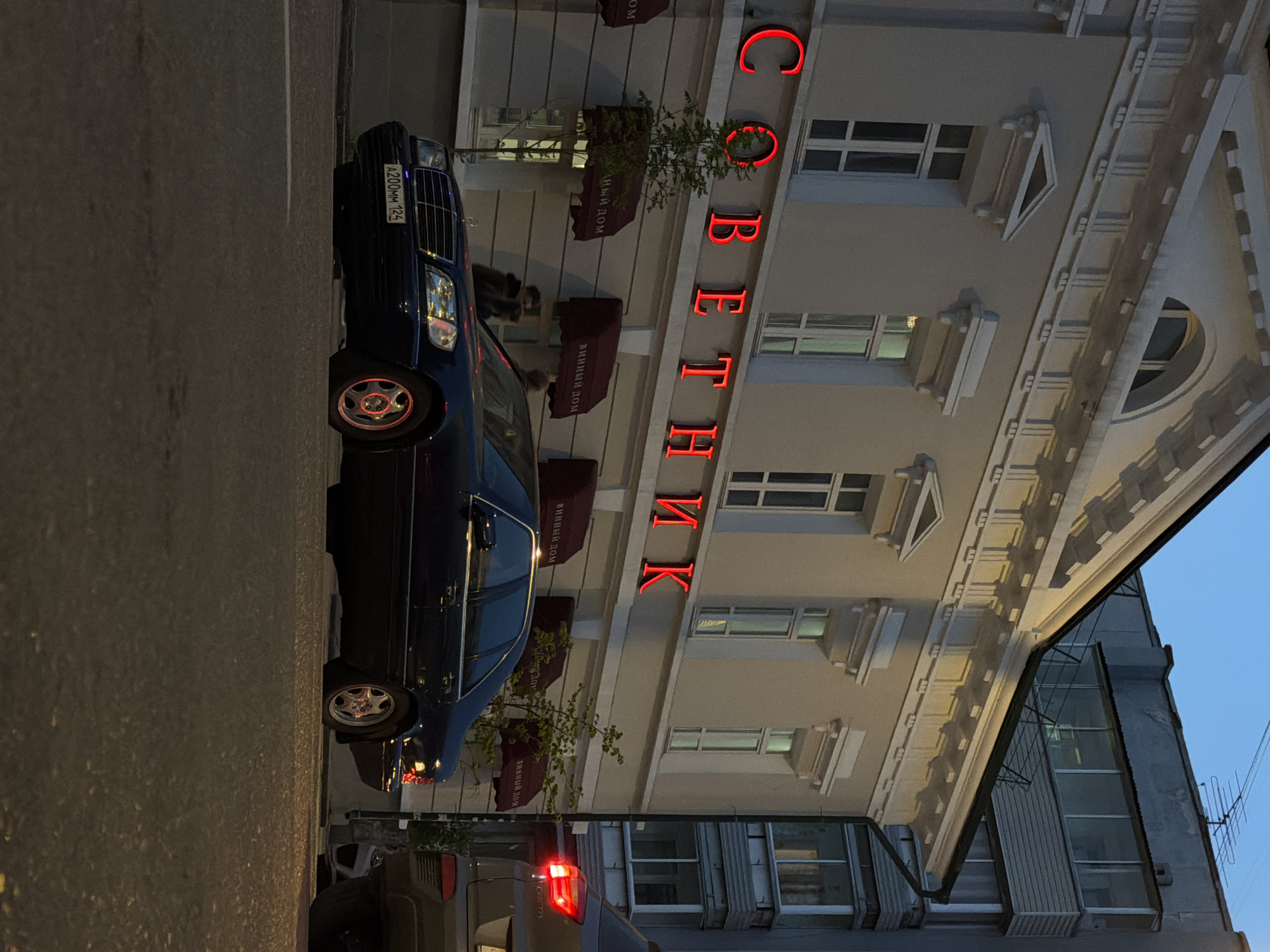 A nighttime street scene in Krasnoyarsk, Russia, presents a dark blue Mercedes-Benz sedan parked on the left side of the road. The vehicle displays license plate A200MM 125. Adjacent to the car stands a light-colored building with windows and maroon awnings. The building's facade prominently features red neon Cyrillic letters spelling "СОВЕТНИК" (SOVETNIK). In the foreground, a portion of a gray vehicle is visible, with its brake lights illuminated. The sky is partially visible in the background, suggesting dusk or early evening.