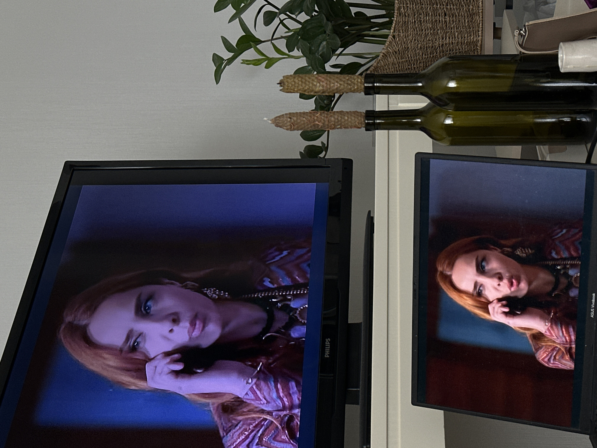 The image captures a domestic scene featuring two screens displaying the same image of a red-haired woman speaking on a phone. The woman wears a patterned top, statement jewelry, and has a concerned expression. The screens appear to be a Philips TV and an ASUS Vivobook laptop. Two green glass wine bottles are on a shelf behind the screens, with decorative straw coverings on their necks. A houseplant and a woven basket are situated above the wine bottles. The scene is indoors, likely in a private residence. There is no information indicating the location is in Tbilisi, Georgia.