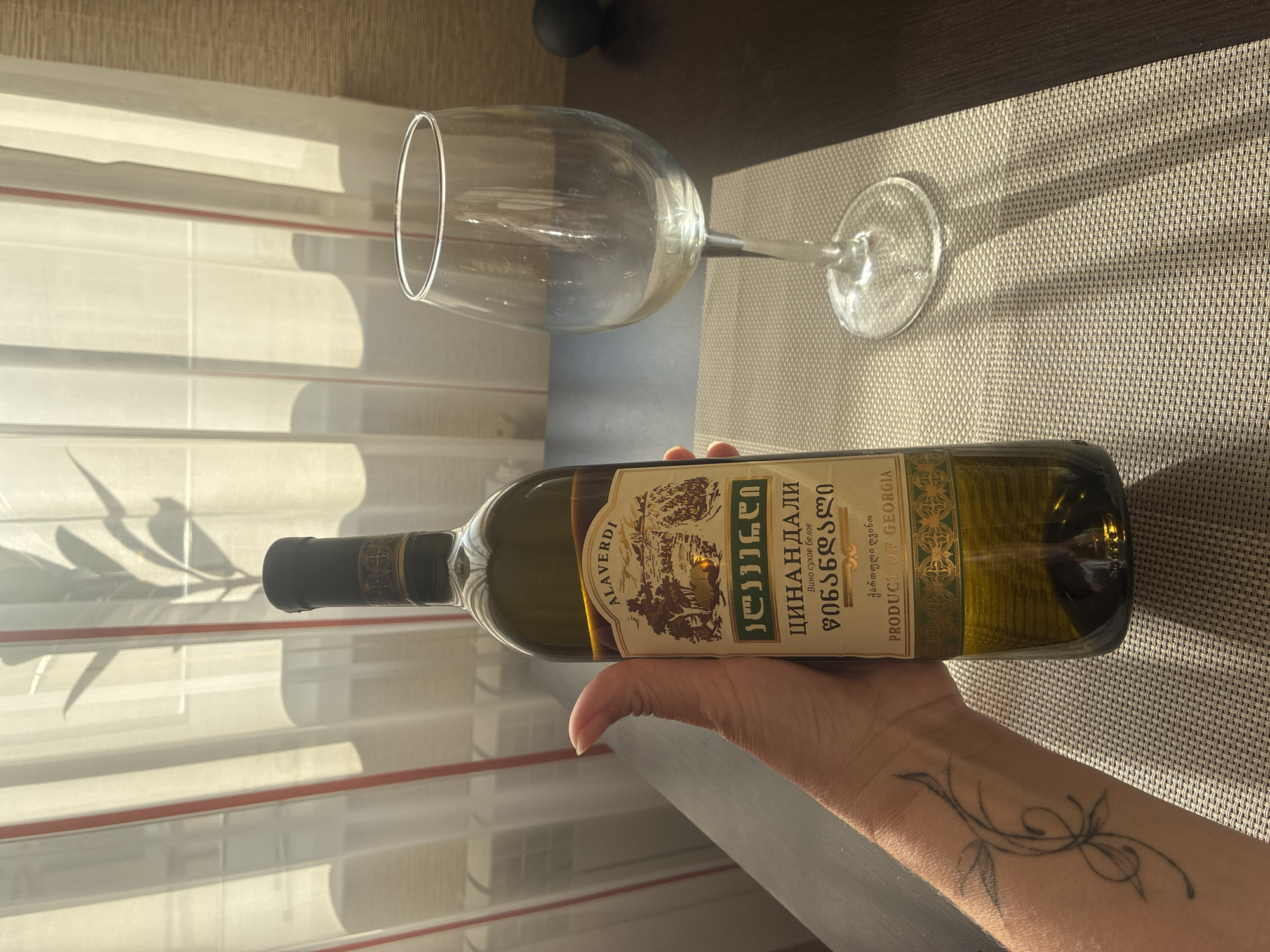 A hand is holding a green glass bottle of Alaverdi Tsinandali white wine. The bottle has a label with text in Georgian and Cyrillic, as well as an illustration of a landscape with trees and a cart. To the right of the bottle is an empty clear wine glass with a stem. The objects are placed on a textured placemat on a dark wooden table. In the background, sheer white curtains with red stripes hang in front of a window, casting shadows on the wall. A plant's shadow is visible on the curtains. The scene appears to be indoors, with natural light filtering through the window. There is no indication of the city of Sochi, Russia.