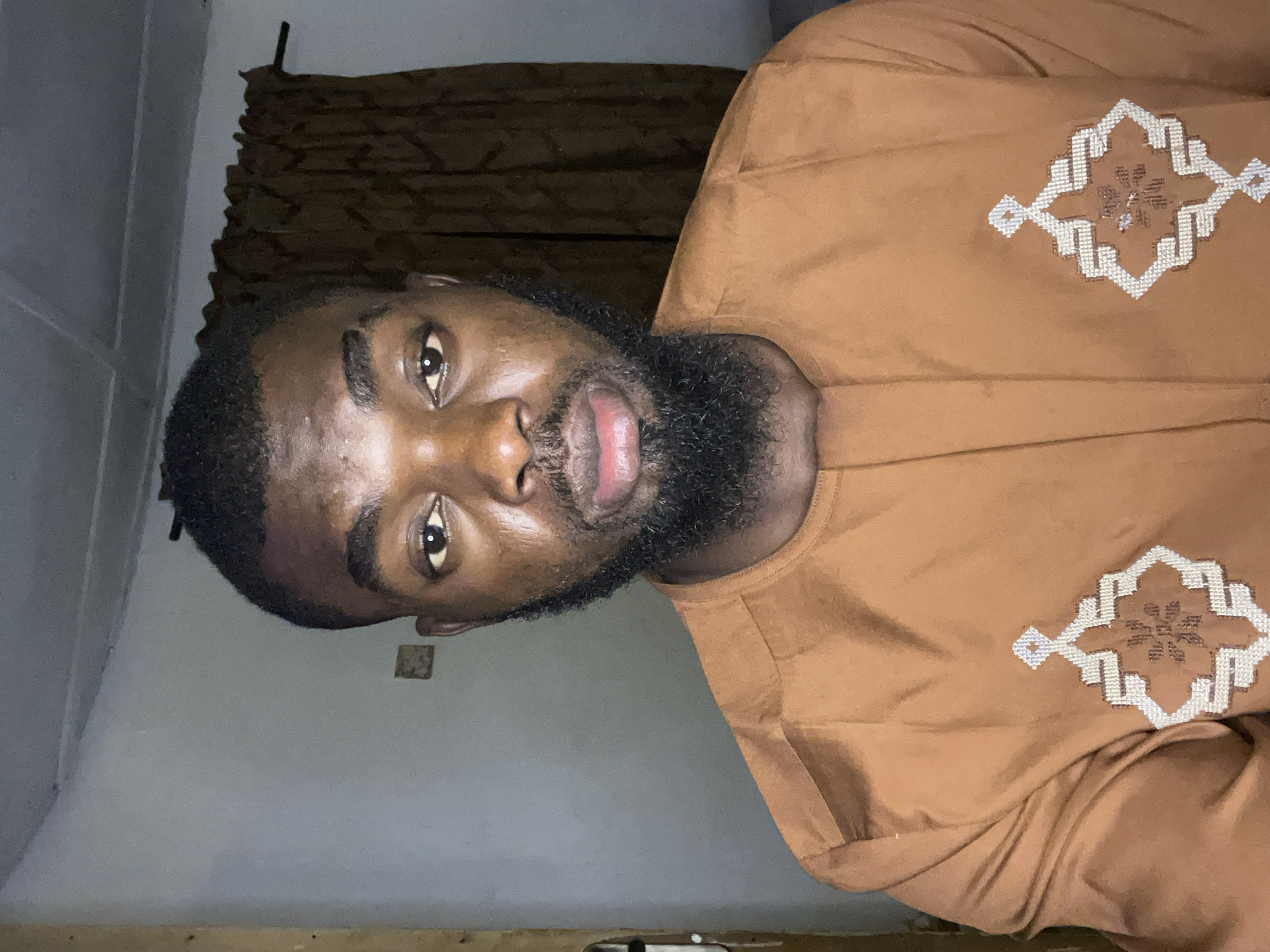 The media file presents a portrait of an adult man, captured indoors. The image is rotated 90 degrees counter-clockwise, showing the man's head on the left and his body extending to the right.
The subject is a dark-skinned man, appearing to be in his 20s or 30s, with short, dark hair and a well-maintained full beard. He has dark eyes and is looking directly into the camera with a neutral to slightly serious expression, making direct eye contact. His lips are slightly parted.
He is wearing a long-sleeved, brown or terracotta-colored garment, possibly a traditional West African tunic. The garment features elaborate, geometric embroidery on the chest area, predominantly in white and light brown or beige threads, forming a symmetrical, star-like pattern within a diamond shape.
The background is an indoor setting with plain, light-colored walls, possibly off-white or light grey. In the upper part of the background, a dark brown curtain or fabric hangs. Towards the bottom, a strip of wood or wooden trim is visible along the base of the wall. There is a small, rectangular, discolored or textured patch on the wall behind the man's head, slightly to his right. The lighting is somewhat dim, suggesting an indoor environment with artificial light, possibly in the evening or night.
The man is posing for the photograph. There are no activities taking place other than the pose itself. No visible text is present in the image. While the location context is provided as Jos, Nigeria, there are no specific visual cues within the image itself to confirm this, beyond the man's appearance and attire which are consistent with the region.