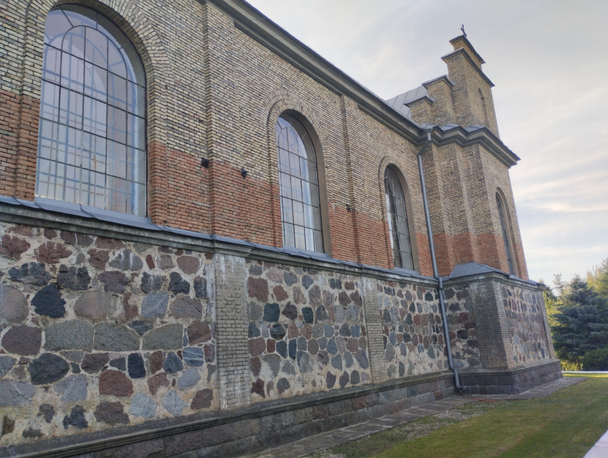 The image depicts the exterior of a brick building, possibly a church, with a stone wall in the foreground. The building's facade features alternating bands of lighter yellow and reddish-brown brickwork. Large arched windows with multiple panes are visible on the upper section of the building. The stone wall is constructed from a mosaic of irregularly shaped stones in various shades of gray, brown, and pink. A grey roofline is visible along the top of the building. A steeple with a cross at its apex rises from the building's corner. The surrounding environment appears to be a grassy area with trees in the background under a cloudy sky. The scene suggests a historical architectural structure in a rural or semi-rural setting, identified as being in the village of Vishnevsky Rural Council, Belarus. There are no people or active events depicted.