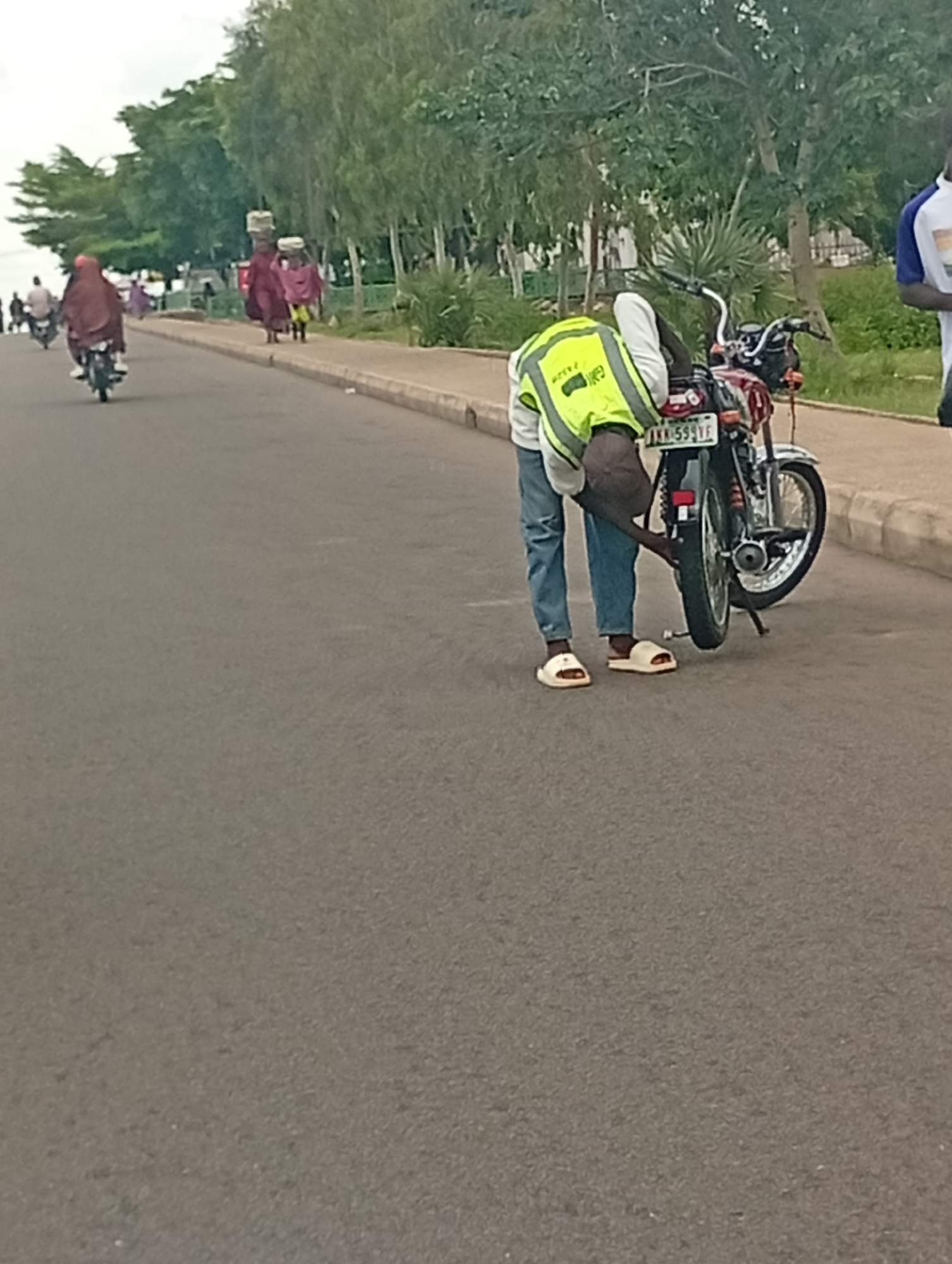 A man wearing a bright yellow reflective vest over a white shirt and blue jeans is bending over a red motorcycle. He appears to be inspecting or adjusting the rear tire of the motorcycle, his right hand near the wheel. He is wearing white slide sandals. The motorcycle has a license plate that reads "AKN-599 VE". In the background, on the right side of the road, another person is visible, wearing a white t-shirt with blue trim and dark shorts.
The scene is on an asphalt road, with a raised concrete sidewalk to the right. Beyond the sidewalk, there are trees and vegetation. In the further background, several people are walking and riding motorcycles. Some individuals in the background are carrying items on their heads, suggesting a marketplace or busy thoroughfare. The sky is overcast. The location is Gombe, Nigeria.