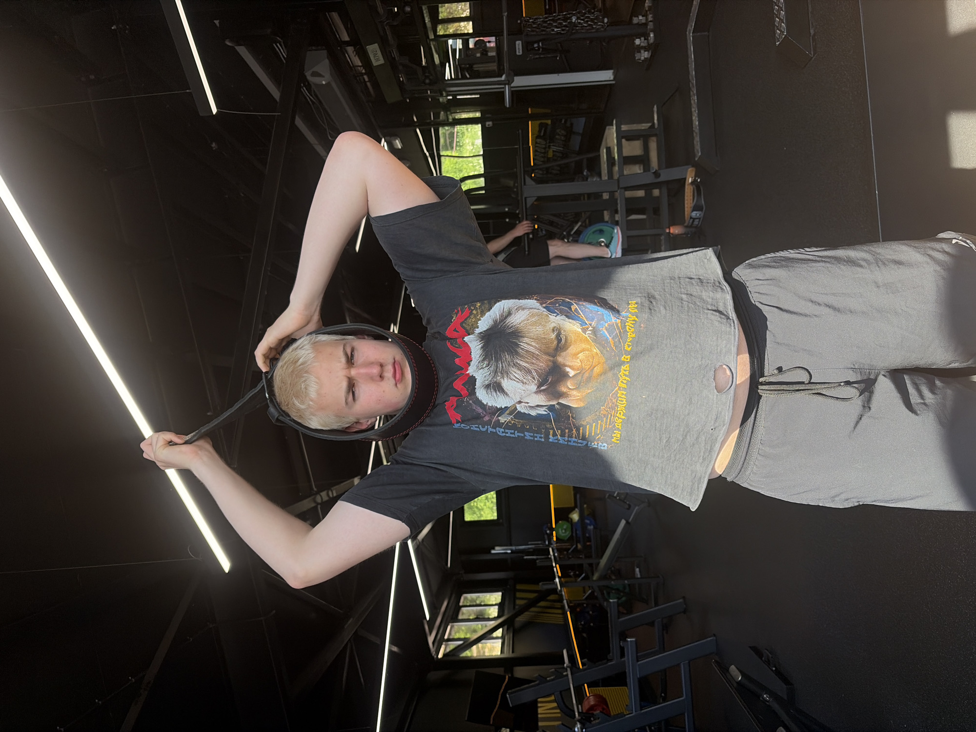A young blonde person with a serious expression stands in a dimly lit gym, holding a black neck harness above their head with both hands. They are wearing a black t-shirt with a graphic print and dark grey athletic shorts. The gym is equipped with various weightlifting machines, benches, and racks. The floor is black rubber, and the ceiling features industrial lighting. Sunlight streams in from windows on the left side of the room, casting shadows. Another person is partially visible in the background, on a weight machine.