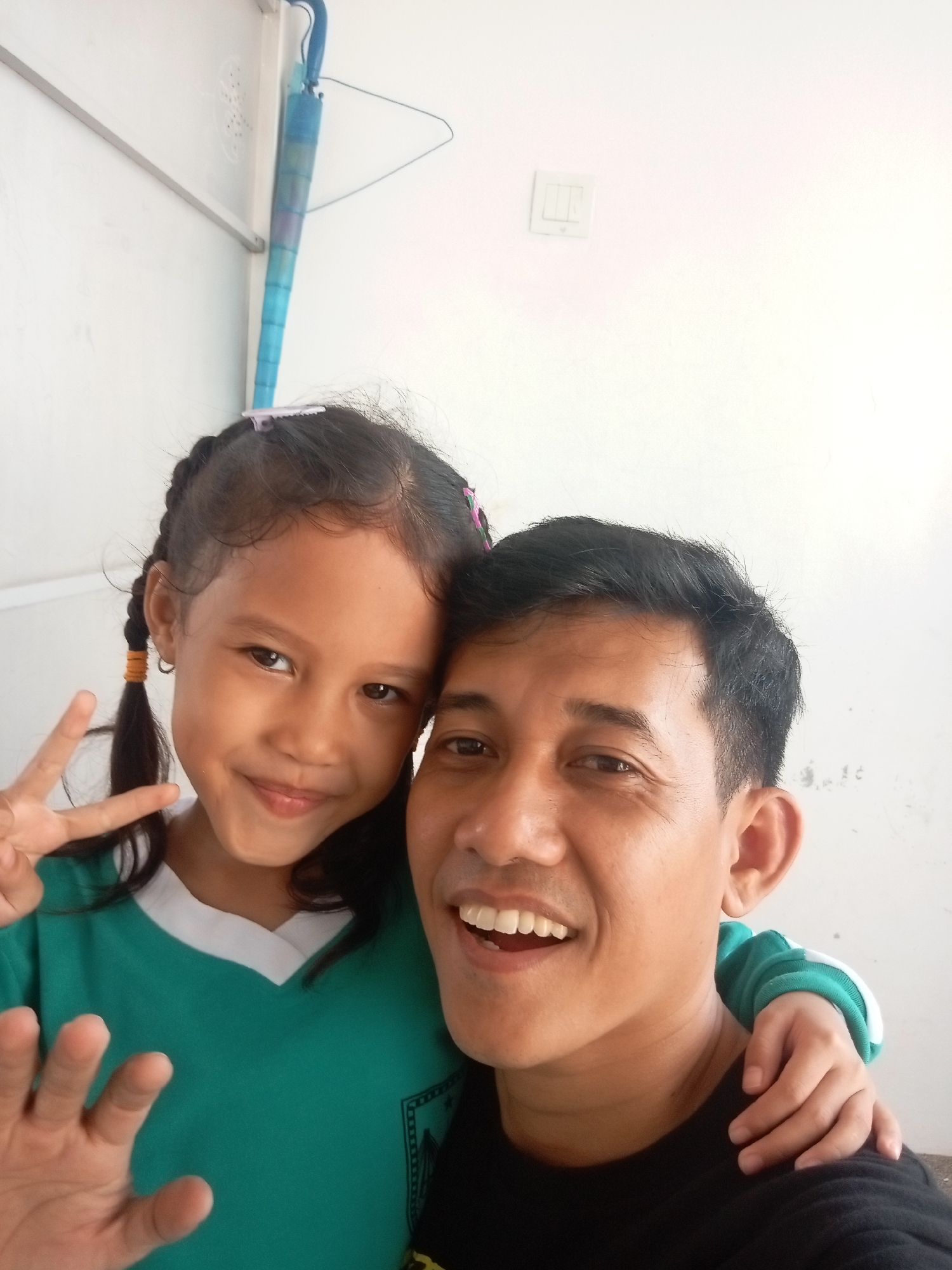 A man and a young girl are posing for a selfie in what appears to be a residential setting in Batam, Indonesia. The man, wearing a black t-shirt, is smiling broadly, showing his teeth. The girl, with braided hair and wearing a turquoise shirt with a white collar, is standing to his left and making a peace sign with her right hand. Her left arm is around the man's shoulders. In the background, a white wall is visible, with a light switch on the right side. An open umbrella, predominantly blue with some purple and darker blue sections, is hanging from a hook on the wall to the left of the subjects. The overall environment suggests an indoor or covered outdoor area, possibly a home or a school in Batam.
