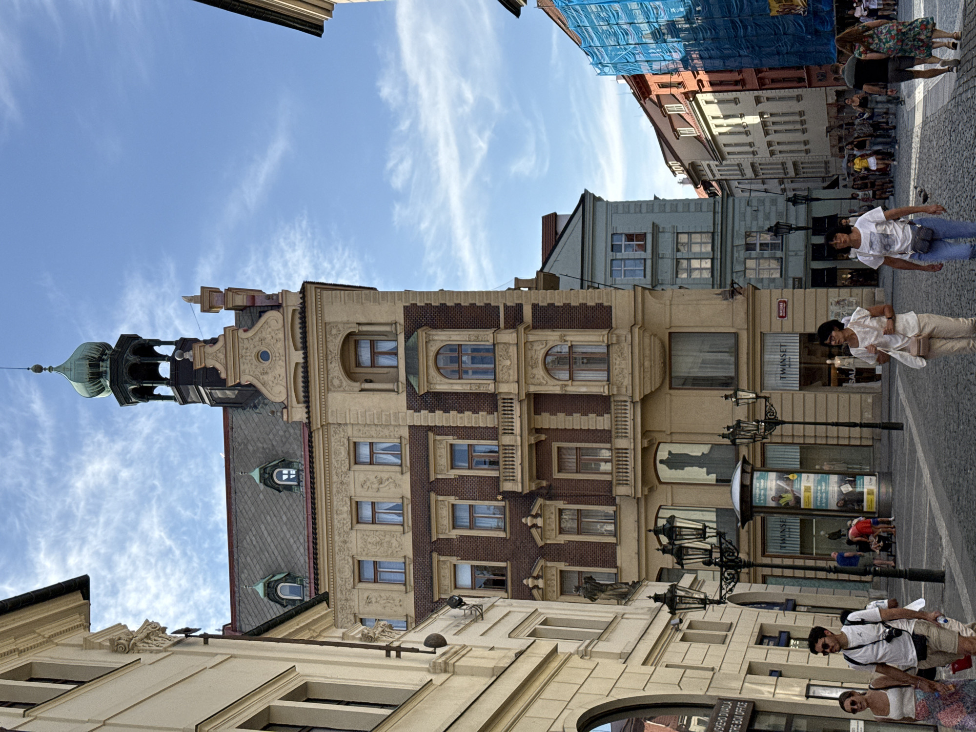 The image depicts a bustling street scene in Prague, Czechia, likely in the Old Town Square area, characterized by its historic architecture and cobblestone streets. The foreground features a couple walking arm-in-arm, the man wearing a white t-shirt, sunglasses, and beige shorts, with a camera around his neck and a crossbody bag. The woman is in a white tank top and a colorful patterned skirt. They are walking on a cobblestone pavement.
Several lamp posts with multiple ornate lights are prominent, particularly to the left of the couple. Behind them, buildings of various architectural styles and colors line the street. A tall, cream-colored building with a red-tiled roof and a green-domed tower dominates the central background. This building displays intricate facade detailing, numerous windows with ornate frames, and balconies. To its left, a lighter-colored building partially obscures the view, showcasing a more neoclassical design.
Further down the street, a cluster of buildings with facades painted in muted tones, including light blue and peach, are visible. On the right side of the street, a building covered in bright blue scaffolding suggests ongoing construction or renovation. Pedestrians are scattered throughout the scene, some walking in groups, others singly, creating a sense of activity and urban life. Shopfronts are visible on the ground floors of some buildings, with signage and displays indicating commercial activity. The sky is bright blue with scattered white clouds, suggesting pleasant weather.