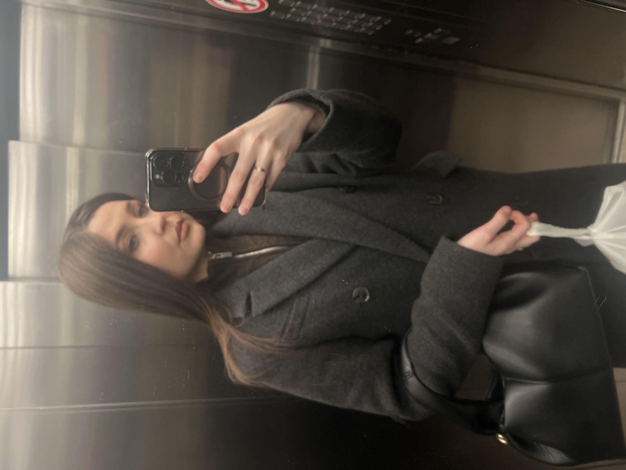The image displays a mirror selfie of a young woman, captured within what appears to be an elevator, evident from the reflective metallic paneling and faint control panel elements. The photo is oriented 90 degrees clockwise from how a person would typically view it.
The subject is a woman with long, dark brown hair, seen from the waist up. She is wearing a dark gray or charcoal-colored double-breasted coat over a brown garment, possibly a sweater or top, visible at her neck. Her left hand is holding a dark-colored smartphone, which features multiple camera lenses and a circular pop socket or grip on its back, indicating she is taking the selfie. A thin, metallic ring is visible on her left ring finger. Her right hand holds a soft, black padded handbag and a crinkled white plastic bag. The woman has a neutral expression as she looks towards the camera reflected in the mirror.
In the reflection behind her, the brushed metal walls of the elevator are prominent. In the upper portion of the reflection, a red circular sign with a diagonal line, commonly indicating "no smoking," is visible. Below this, part of an elevator control panel with blurry buttons can be faintly discerned. The lighting is artificial, consistent with an indoor environment.