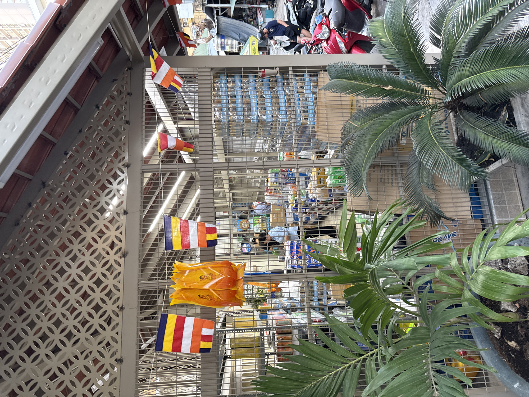 The image depicts a scene in Nha Trang, Vietnam, likely at a shop or market stall. In the foreground, two potted plants, one a palm and the other a cycad, are visible. Behind them, a metal fence separates the viewer from the interior. Hanging from the fence are several Buddhist prayer flags. The interior of the stall is filled with shelves stocked with various goods, including what appear to be canned drinks and other packaged items. Two people are visible inside the stall, one standing and another interacting with shelves. Outside the fence, to the right, several motorcycles are parked, and a man wearing a helmet is seated on one. The overall environment suggests an outdoor commercial setting within a Vietnamese city, characterized by its typical street life and retail practices.