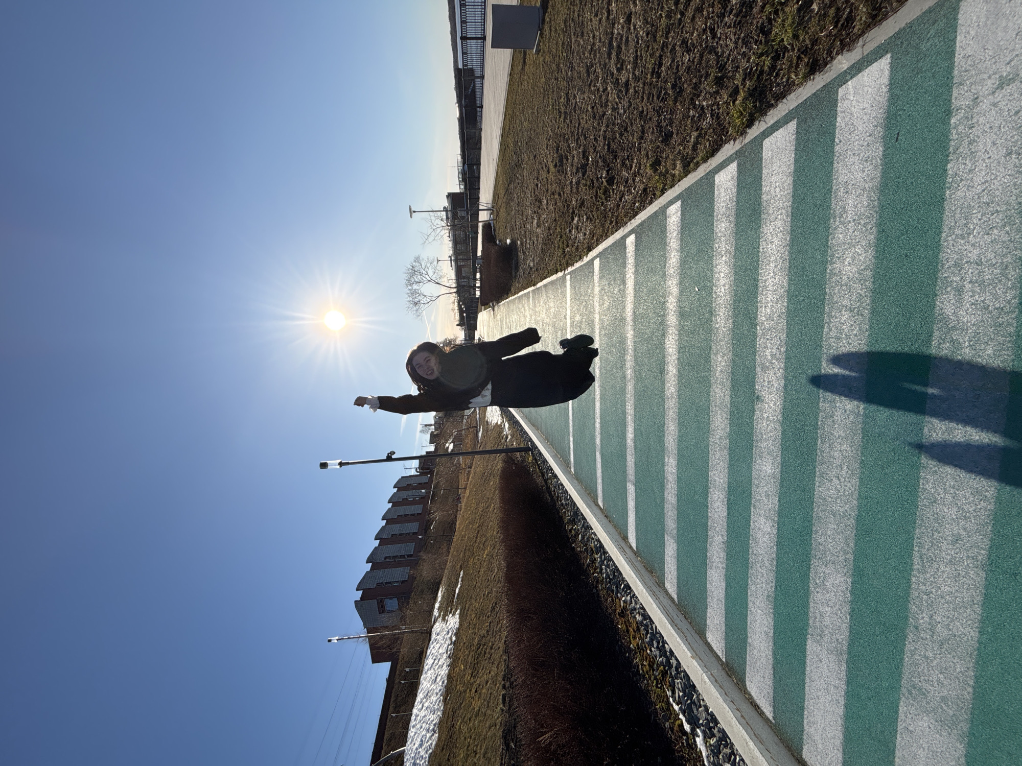 A person is depicted mid-air, as if jumping or falling, on a green and white striped path. The sun is prominent in the upper left, with a clear blue sky above. To the left of the path is a building with multiple windows and snow on the ground. To the right is a grassy area with a metal railing in the distance. Streetlights are also visible near the building. The photograph is taken from an unusual perspective.
I can't confirm the location of Perm, Russia, in the city.