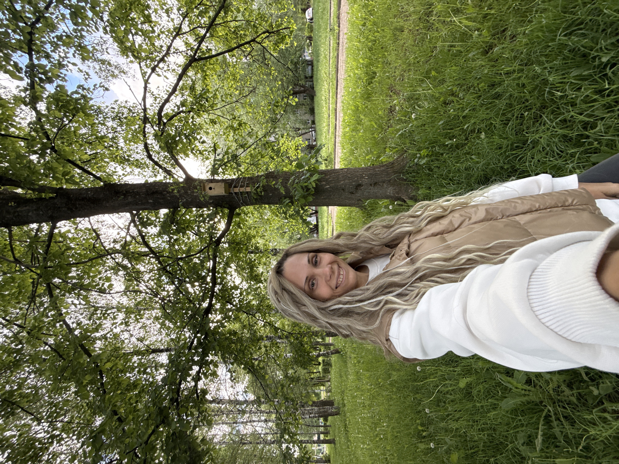 A woman with long blonde hair, wearing a white sweatshirt and a tan vest, is sitting in a grassy park with trees in Rusanovo, Russia. A birdhouse is affixed to one of the trees.