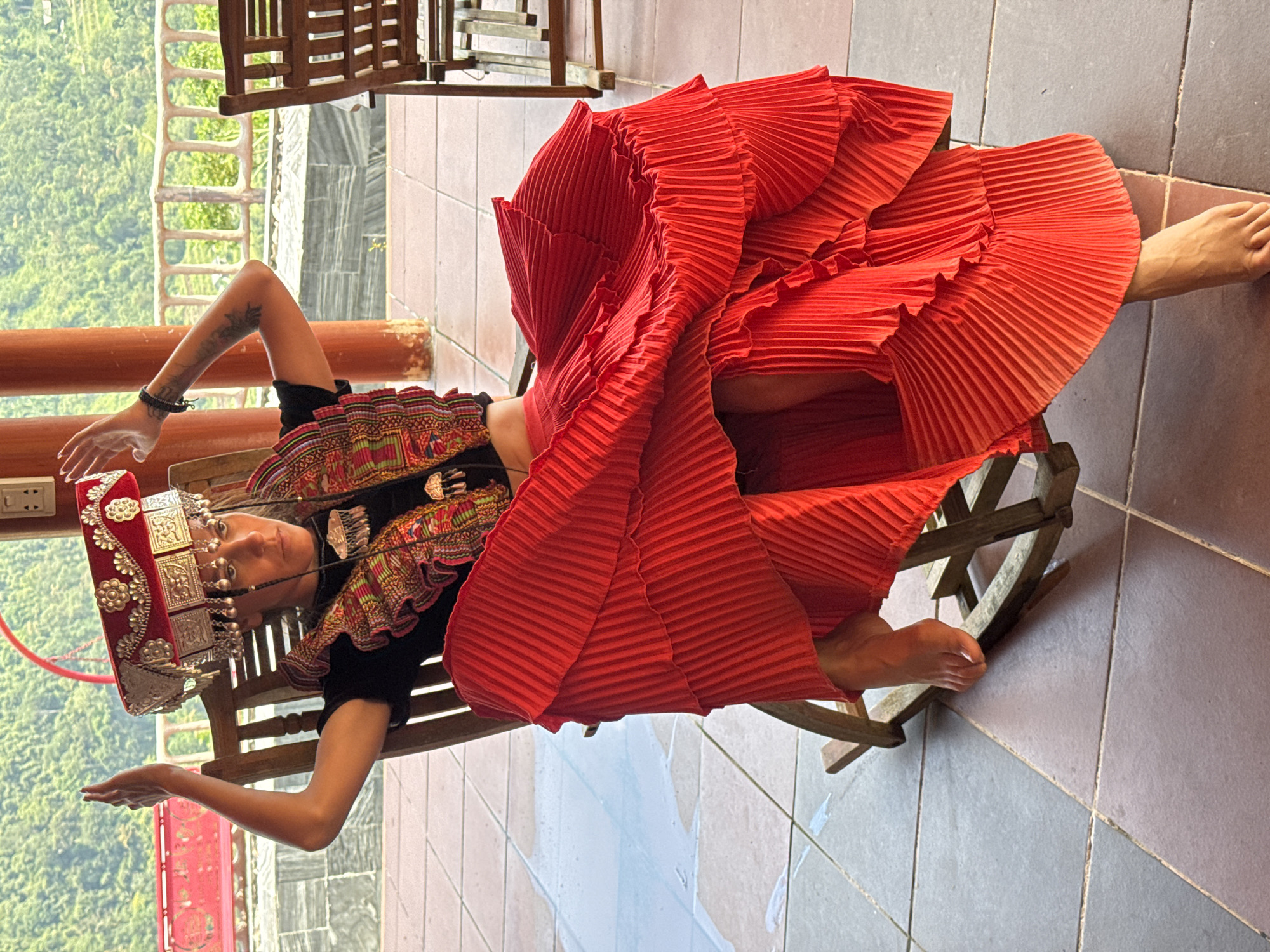 A woman poses in elaborate traditional attire, seemingly lying back on a wooden rocking chair on a tiled outdoor or semi-outdoor balcony. The image is rotated sideways. She wears a tall, red fabric headdress richly adorned with silver metal plates and dangling chains. Her black top features intricate, colorful embroidery with geometric and floral patterns. A voluminous, bright red pleated skirt flares around her, obscuring most of the chair.
The woman is barefoot, her arms are bare, revealing tattoos on her left arm. She has a serene expression, looking towards the camera. Her hands are open and extended slightly. The background features a wooden railing and a view of lush green mountains, suggesting a natural and elevated location. The bright lighting indicates daytime and clear weather. The floor is covered with light brown and grey tiles. There is no visible text in the image. The attire, combined with the Thông Nguyên Commune, Vietnam context, suggests traditional dress of a local ethnic group.