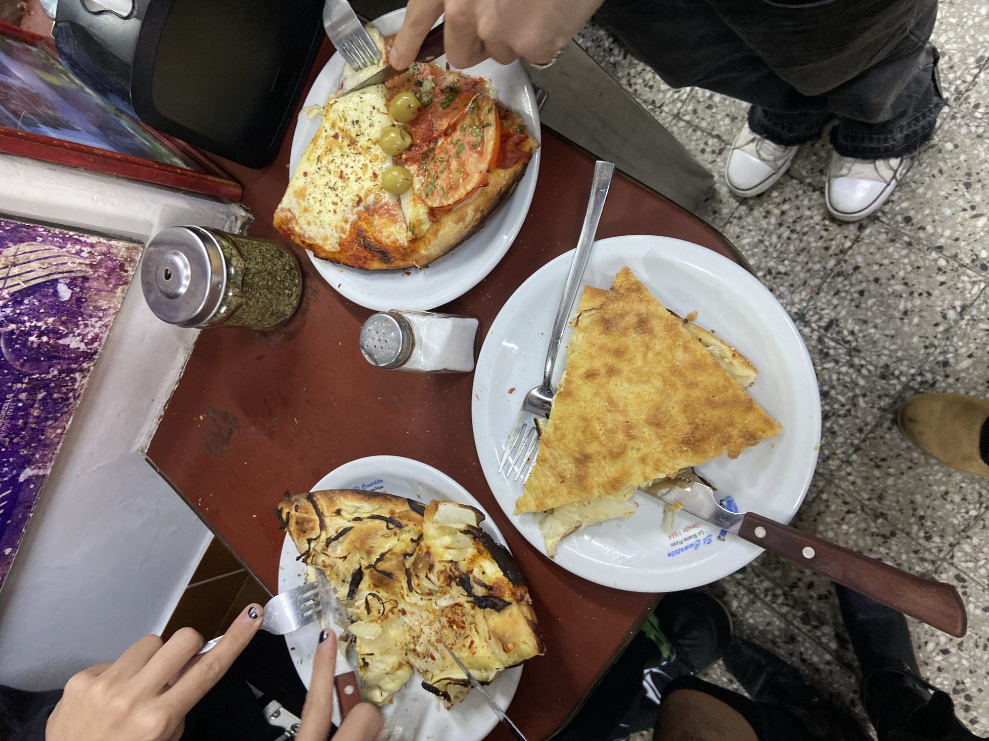 The image shows a table at El Cuartito, a pizzeria in Buenos Aires, Argentina. Three plates of different kinds of pizza are on the table. The pizza on the upper left plate has cheese, tomato, and green olives. The pizza on the middle right plate is plain cheese pizza. The pizza on the bottom left plate has onions. Several hands are visible, some holding forks and knives to cut the pizza. A jar of herbs and a salt shaker are also on the table. The floor has a speckled tile pattern. People's feet are visible around the table.