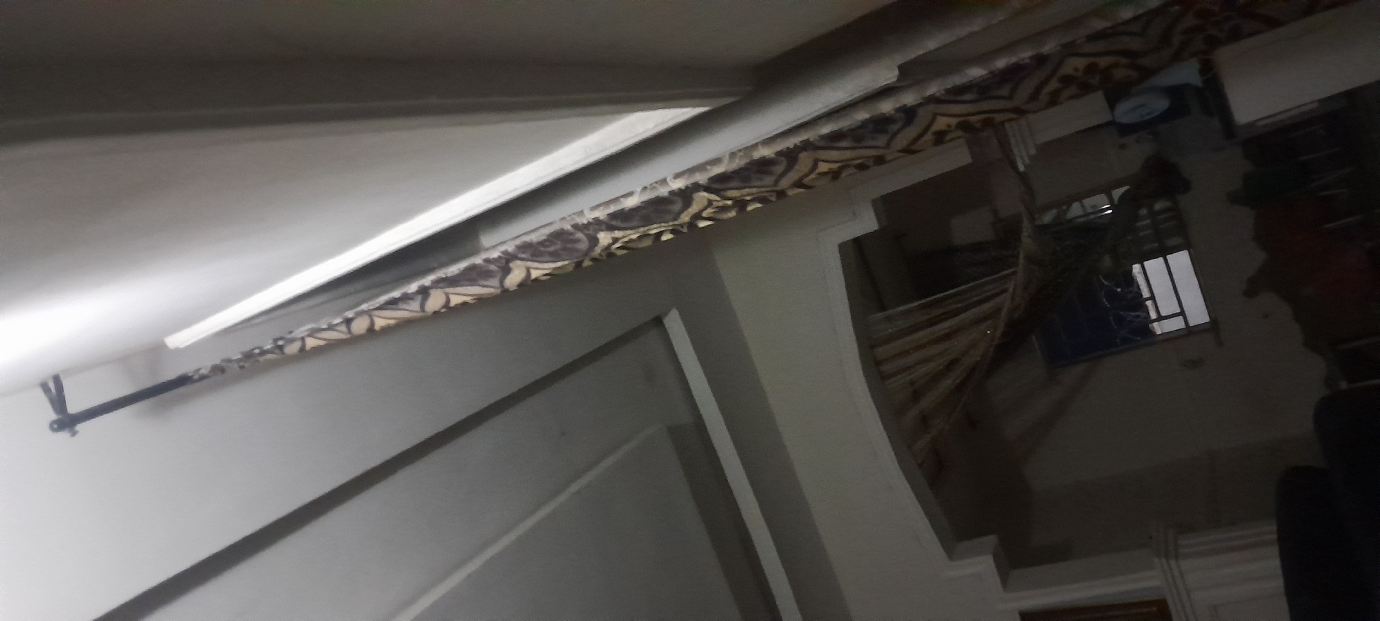 A black curtain rod extends diagonally from the top left to the middle right of the frame, with a dark fabric curtain hanging from it. The curtain has a repeating pattern of beige and dark brown floral or paisley designs. The background consists of a white ceiling with recessed lighting or architectural details. Below the ceiling, an arched opening reveals a dimly lit room. Through the arch, a window with metal bars is visible. The lower part of the image shows a dark, plush chair or sofa in the foreground. The scene is dimly lit, with the primary light source appearing to be from the ceiling and possibly the window. The overall impression is of an indoor residential setting.