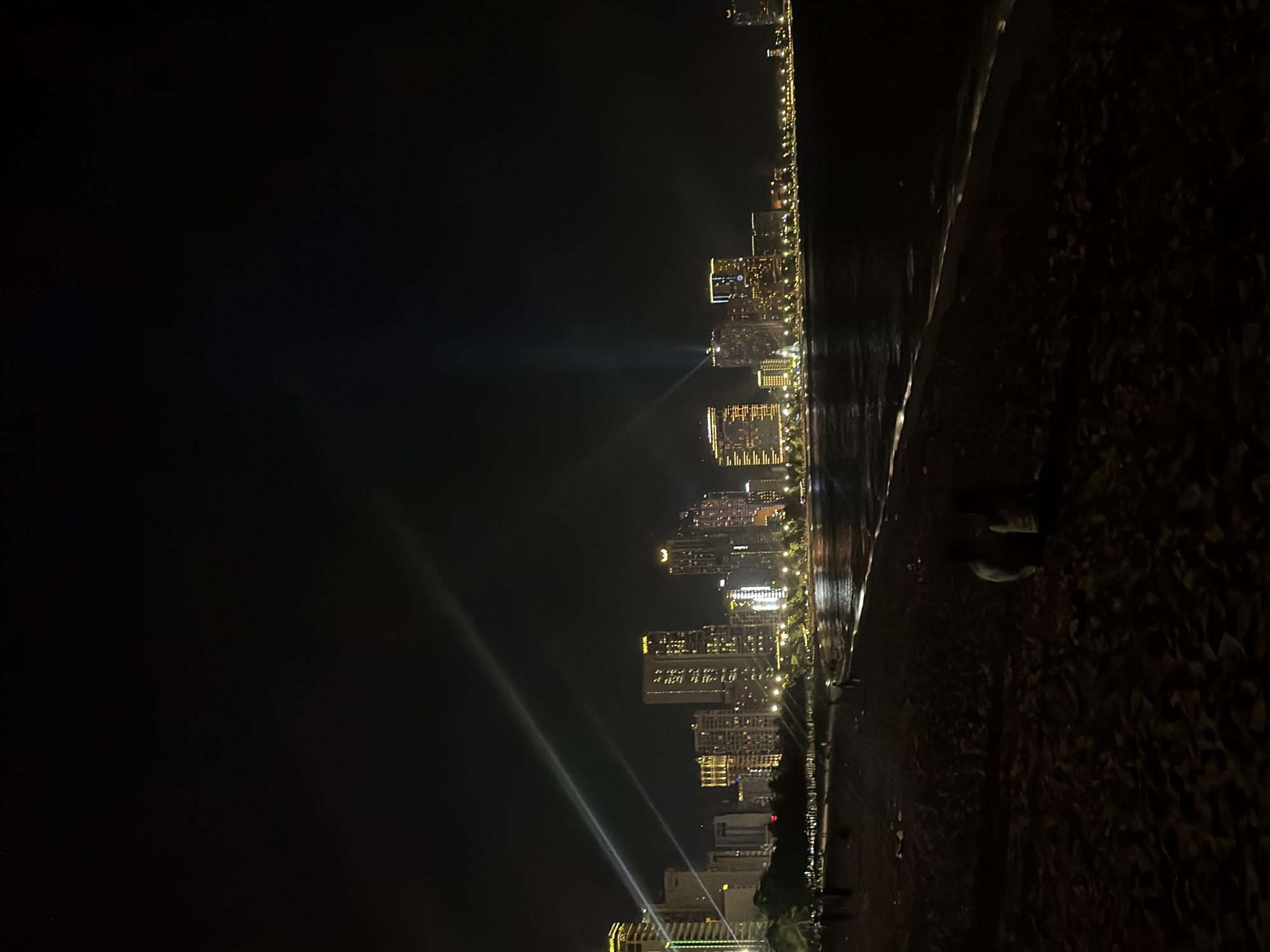 The image shows a nighttime view of the coastline in Nha Trang Ward, Vietnam. The shoreline runs vertically through the right side of the frame with a dark sandy or pebbly beach in the immediate foreground.
Along the coast, there is a series of buildings that include high-rise hotels and apartments. The buildings are illuminated with multiple lights. Some of the buildings have spotlights that shine up into the night sky.
The sea or ocean is adjacent to the shoreline and reflects the lights from the buildings. The background is predominantly dark, with some lights visible farther along the coastline, suggesting the continuation of the city. No individuals are distinctly visible.