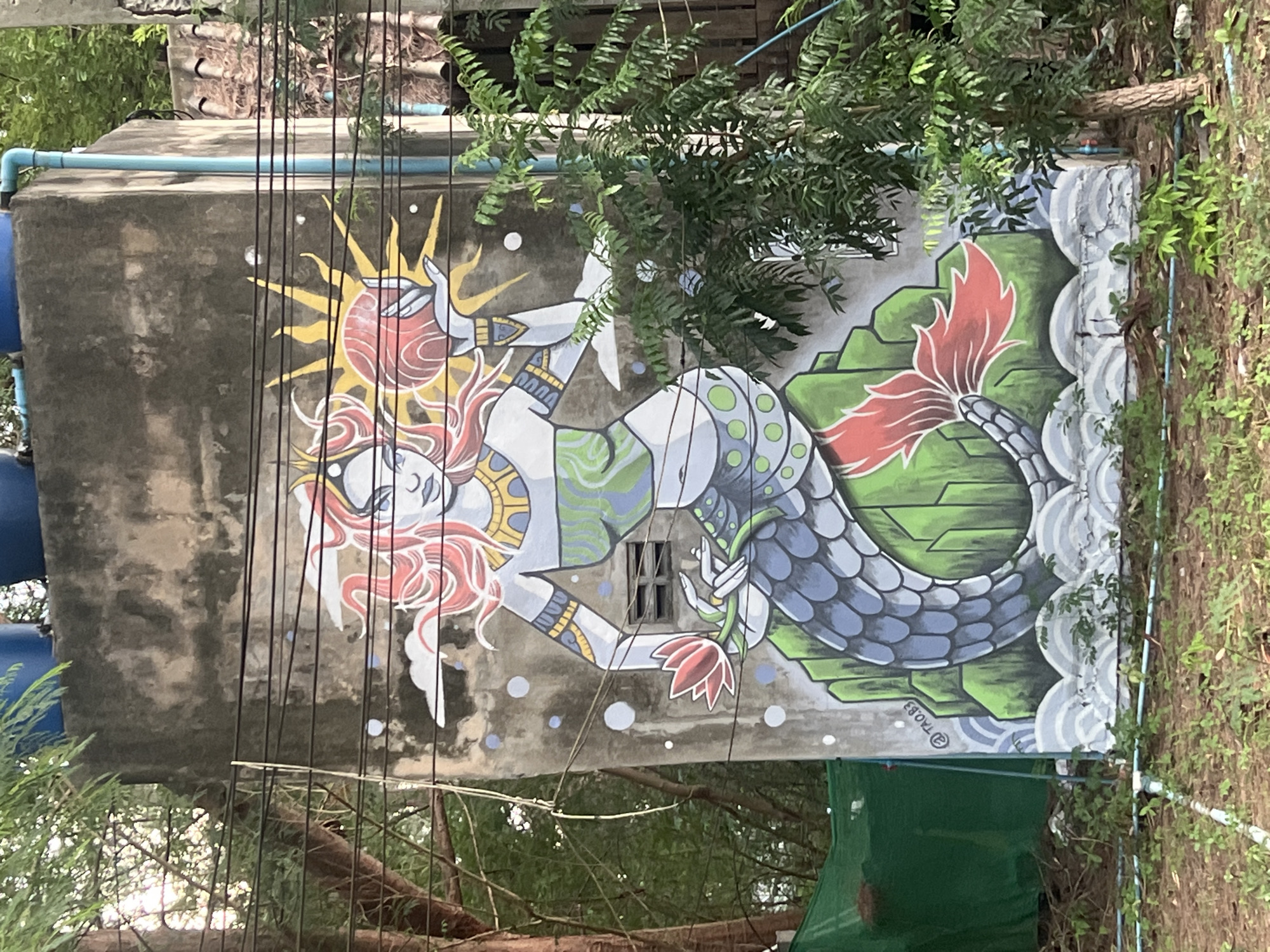 An outdoor mural is painted on a weathered concrete wall in Ko Pha-ngan District, Thailand. The artwork depicts a mythical mermaid with flowing red hair and ethereal features, adorned with gold jewelry and a green patterned torso. She is positioned on top of a cluster of green rocks, clutching a red lotus flower in her left hand. In her right hand, she holds a glowing red orb, possibly a sun, with yellow rays emanating from it. The mermaid's tail is a large, scaled, grey fish tail with red fins. The mural is surrounded by lush green foliage and trees, and overhead, electrical wires crisscross the scene. The base of the wall is marked with what appears to be a signature "@TAO.83".