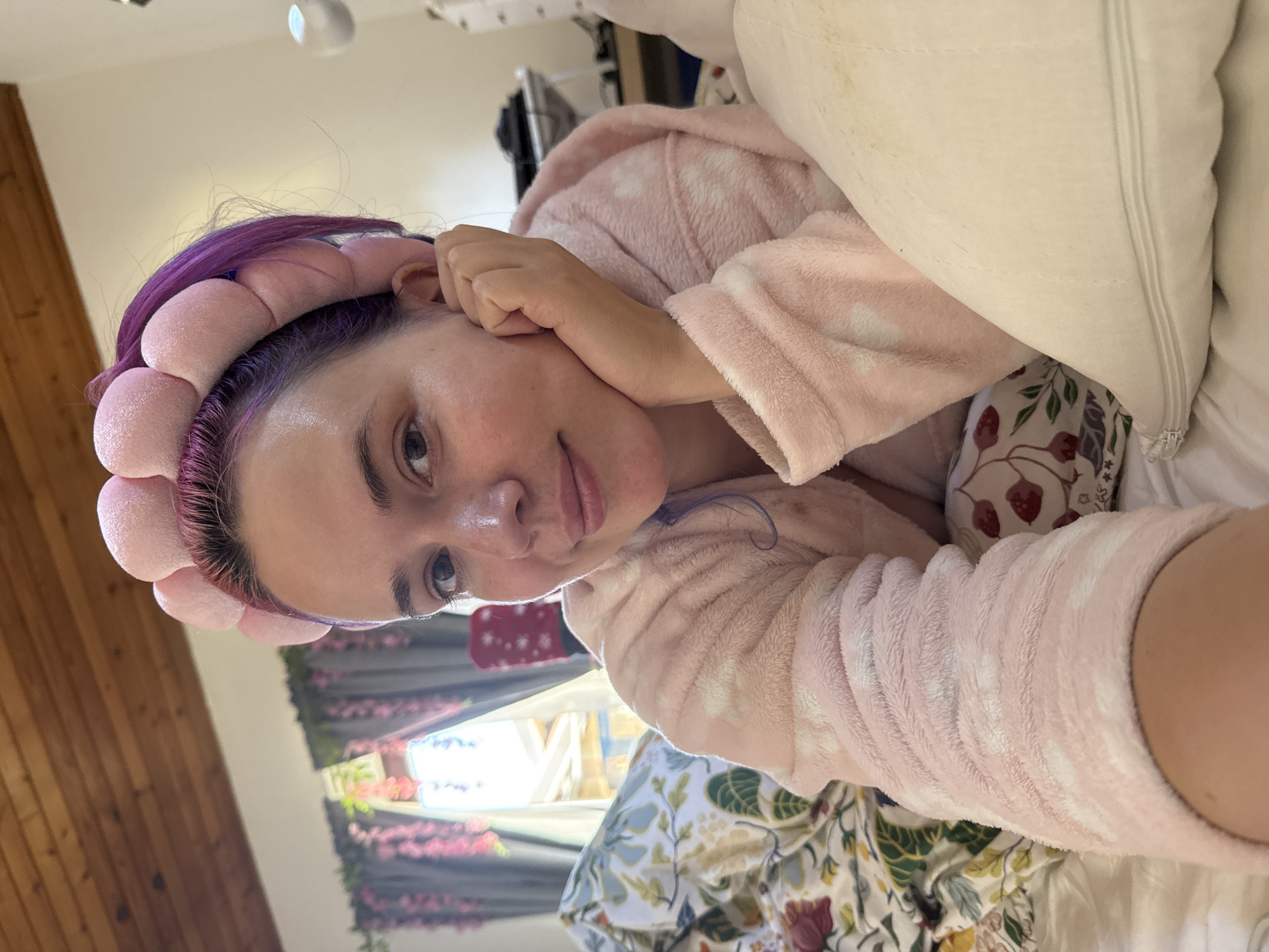 The image shows a person with purple hair wearing a pink, bubble-like headband and a pink bathrobe with white polka dots. The person is lying down on a bed with a floral-patterned duvet and a white pillow. The person is facing the camera and resting their hand near their face. Behind the bed, partially visible, is a window with floral curtains. There is no evidence in the image to determine if the location is in the city of City of Zagreb, Croatia.
