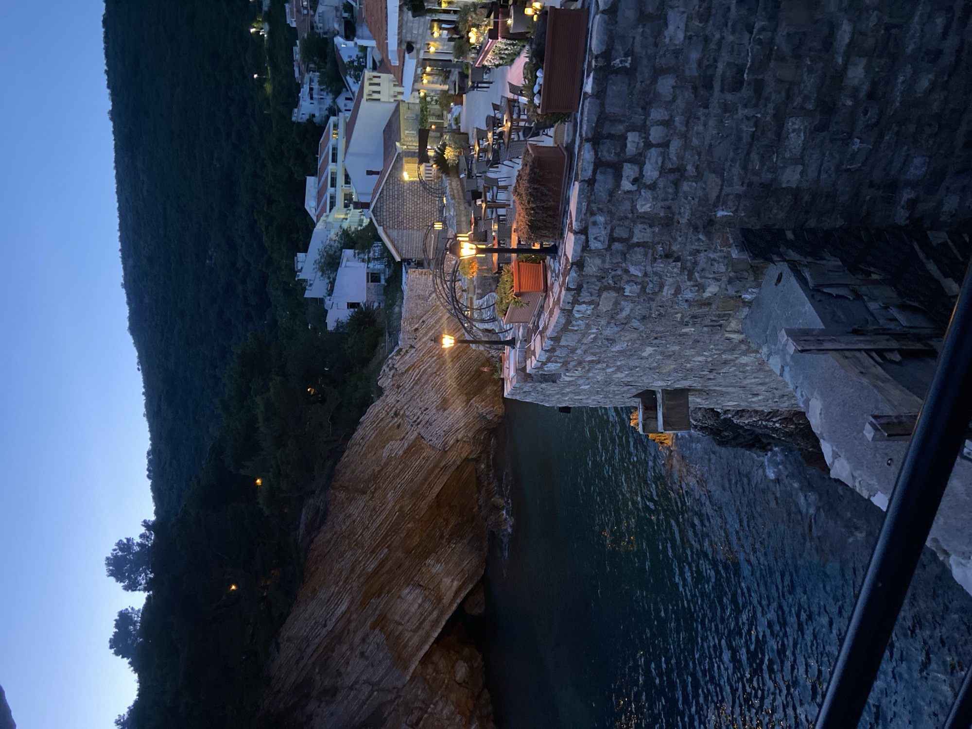 The image shows a nighttime scene in Petrovac centar, Montenegro. A steep, densely wooded hillside descends to meet the sea, where a rocky outcrop juts into the water. Along the upper edge of the outcrop, a stone wall with decorative street lamps defines a terrace area. The terrace is furnished with tables and chairs, suggesting an outdoor restaurant or cafe. Further back, white buildings with red-tiled roofs are visible, partially illuminated by artificial lights. Below, dark water fills the scene, reflecting the light from the cliff and buildings.