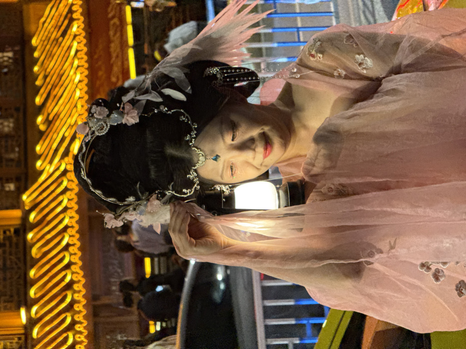 An East Asian woman, approximately middle-aged, is depicted at night in Yuzhong District, China. She wears an elaborate traditional Chinese costume, featuring a sheer, light pink gown with embroidered floral patterns. Her dark hair is styled in a high, ornate updo, embellished with silver-toned hairpieces, pearls, light pink fabric flowers, and pale feathers. A silver-toned forehead ornament with a blue teardrop jewel is visible, and she has red lipstick. She holds a small, black-framed lantern emitting a bright white light in her right hand, which is draped with the sheer fabric of her sleeve, and gazes subtly downward towards it. The background features a blurry street scene with a metal pedestrian barrier, a dark vehicle, and indistinct figures, set against a building displaying prominent golden, wavy illuminated roofline ornamentation, indicative of traditional Chinese architecture.