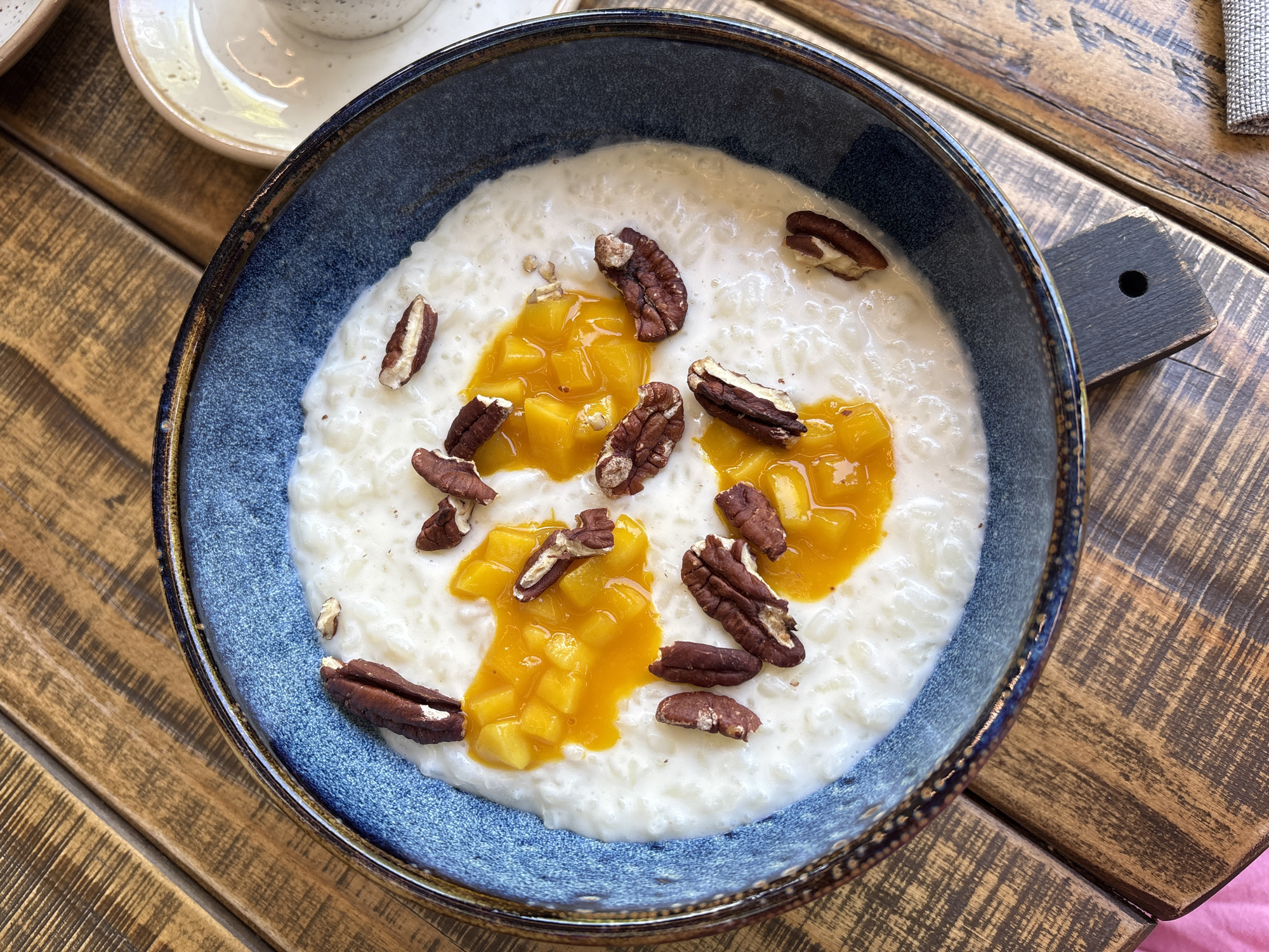A single, overhead shot depicts a serving of a creamy, white rice-based dish, likely rice pudding or kheer, presented in a dark blue ceramic bowl with a rustic, irregular brownish-gold rim. The dish is garnished with multiple whole and broken pecan halves, scattered across the surface, and two distinct dollops of a vibrant orange-yellow fruit compote or puree, containing visible diced fruit pieces, possibly mango or apricot.
The bowl rests on a rustic wooden table composed of multiple planks exhibiting prominent wood grain, varying shades of brown, and distressed markings. Partially visible elements in the background include: the rim of a speckled, light-colored ceramic cup or small bowl to the upper right; a dark, rectangular wooden or slate object with a circular hole to the lower right, positioned next to a folded, light-colored woven fabric item, possibly a napkin. A small section of a bright pink object, likely fabric, is visible in the lower-left corner.
No individuals are present in the image, and no specific actions are occurring. The environment appears to be an indoor setting, possibly a cafe or restaurant, illuminated by ambient light. The specific location in the city of Krasnoyarsk, Russia, cannot be determined from the visual information provided in the image.