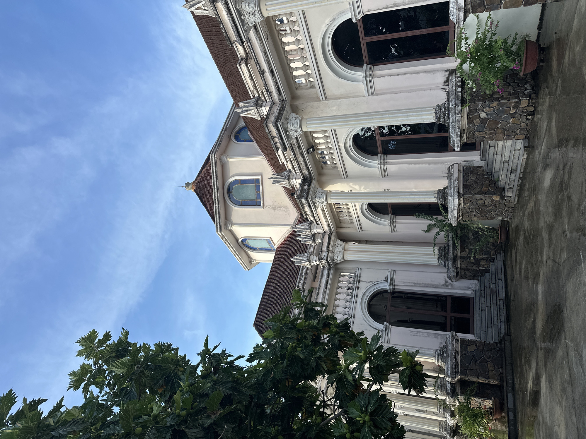 This image shows a historic building in Diên Khánh, Vietnam, likely a church or a similar religious structure, with a light-colored facade and a reddish-brown tiled roof. The building features ornate architectural details, including arched windows with stained glass, classical columns, decorative cornices, and a domed central section. The exterior is adorned with stone accents at the base of the columns and around the entrance stairs. Lush green trees with large leaves partially obscure the left side of the building, and potted plants are visible near the entrances, suggesting a well-maintained garden area. The ground in front of the building appears wet, possibly from recent rain, and reflects the surrounding structures. The sky above is a clear blue with a few wispy clouds. No people are visible in the scene. The perspective is low-angle, emphasizing the grandeur of the architecture against the sky.