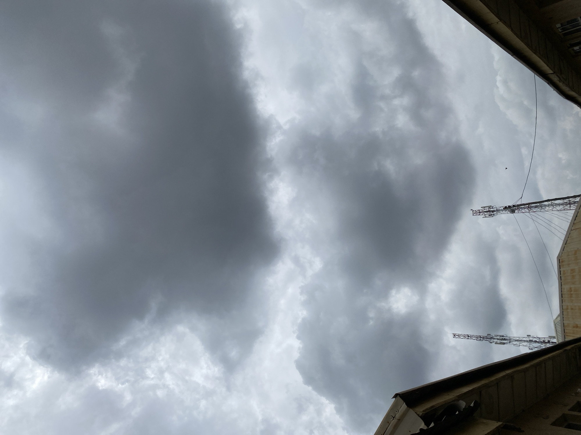 An upward-facing, medium shot captures a dramatic, overcast sky filled with dense, gray clouds. Two tall, lattice-like telecommunication towers are visible, one to the left and one to the right of center, with wires connecting them. The roofs and upper sections of two buildings flank the bottom corners of the frame, their eaves angled towards the sky. A small bird is visible in flight against the cloudy backdrop between the two towers.