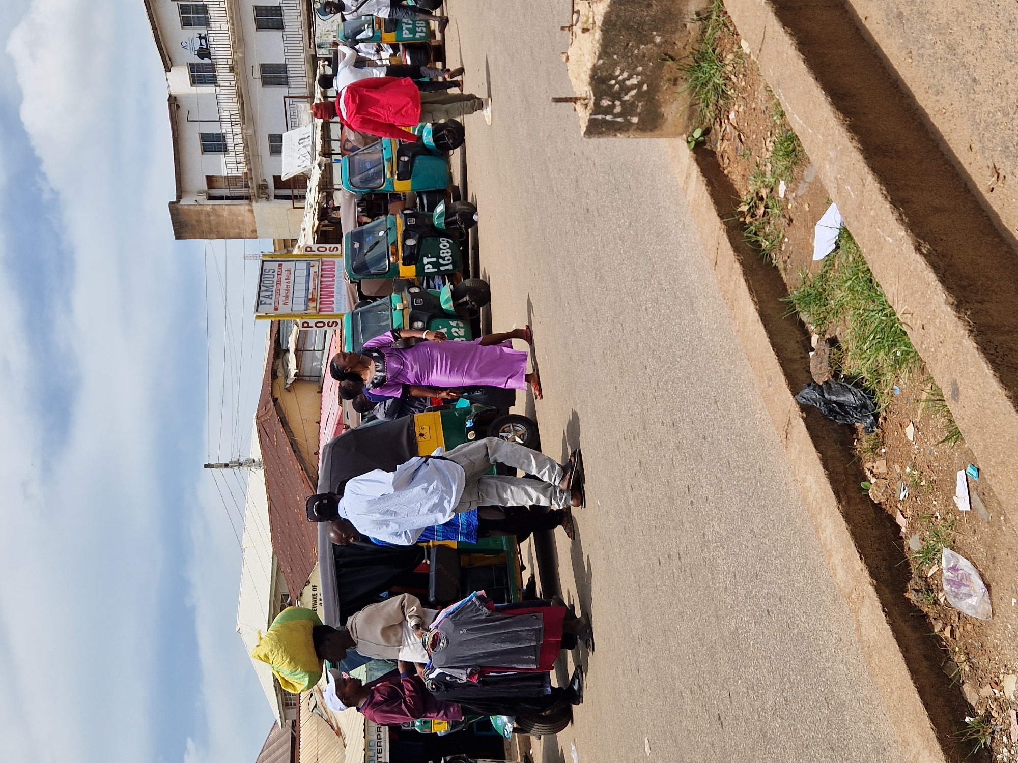 The image captures a busy street scene in Jos, Nigeria, likely a commercial area. The foreground features a paved road with a concrete curb and a narrow, overgrown drainage ditch containing scattered debris, including plastic bags and paper.
In the mid-ground, a row of auto-rickshaws, painted in vibrant green and yellow, are parked along the side of the road, suggesting they are available for hire or are part of a transportation hub. A sign for "FAMOUS WHOLESALES & RETAILS" and "DOWNLOAD MUSIC, AUDIO & VIDEO" is visible in the background, indicating the presence of businesses.
Several individuals are present. On the left, two men are standing near an auto-rickshaw; one is wearing a maroon shirt and a white cap, while the other wears a light brown hoodie and is carrying a bundle of clothes on a hanger. A third man, in a white shirt and grey pants, stands behind them, facing away from the camera. Further to the right, a woman in a long purple dress is walking towards the auto-rickshaws. Another man in a red jacket and cap is seen walking away from the camera towards the auto-rickshaws, and behind him, another man in a white shirt is also visible. The architecture in the background consists of multi-story buildings with balconies and corrugated metal roofs, typical of urban settings in Nigeria. The sky is partly cloudy, suggesting it is daytime. The overall atmosphere is one of commercial activity and daily life in a Nigerian city.
