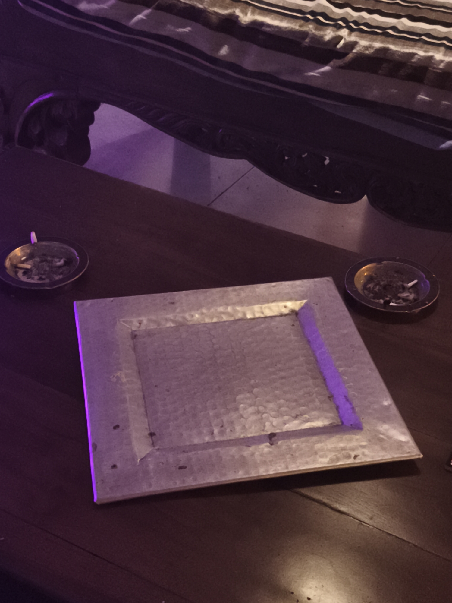 The image shows a dimly lit, dark brown table with a hammered metal serving tray in the center. To the left and right of the tray are ashtrays containing cigarette butts and ash. The background features a bed with striped bedding and carved wood details. The specific location in Barcelona, Spain, cannot be determined from the image.