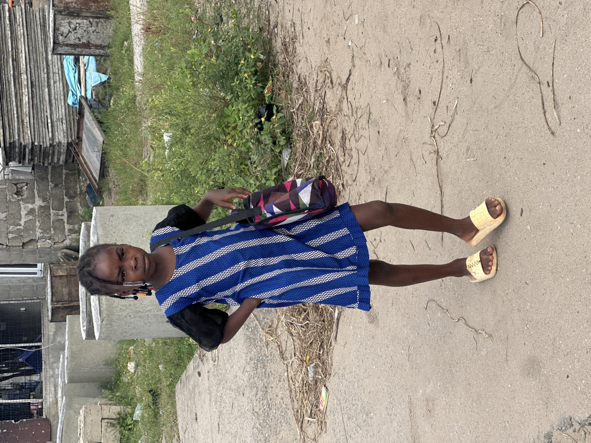 A young Black girl, approximately seven years old, stands on a dirt and asphalt path in Badagry, Nigeria. She is facing the camera, with her left hand on her hip and her right arm bent, her hand near her side. She has dark skin, black braided hair with colorful beads at the ends, and is wearing a blue and white striped dress with black puffed sleeves and yellow slide sandals. A patterned messenger bag with geometric shapes in pink, purple, blue, and white is slung over her right shoulder.
The background includes a partially constructed building with grey cinder blocks and a metal fence, stacks of grey corrugated metal sheets, and green vegetation with small yellow flowers. Scattered debris, including dry leaves, twigs, and plastic, litters the ground around the girl and in the background. The overall scene depicts an outdoor, somewhat unkempt environment in what appears to be a developing area of the city.