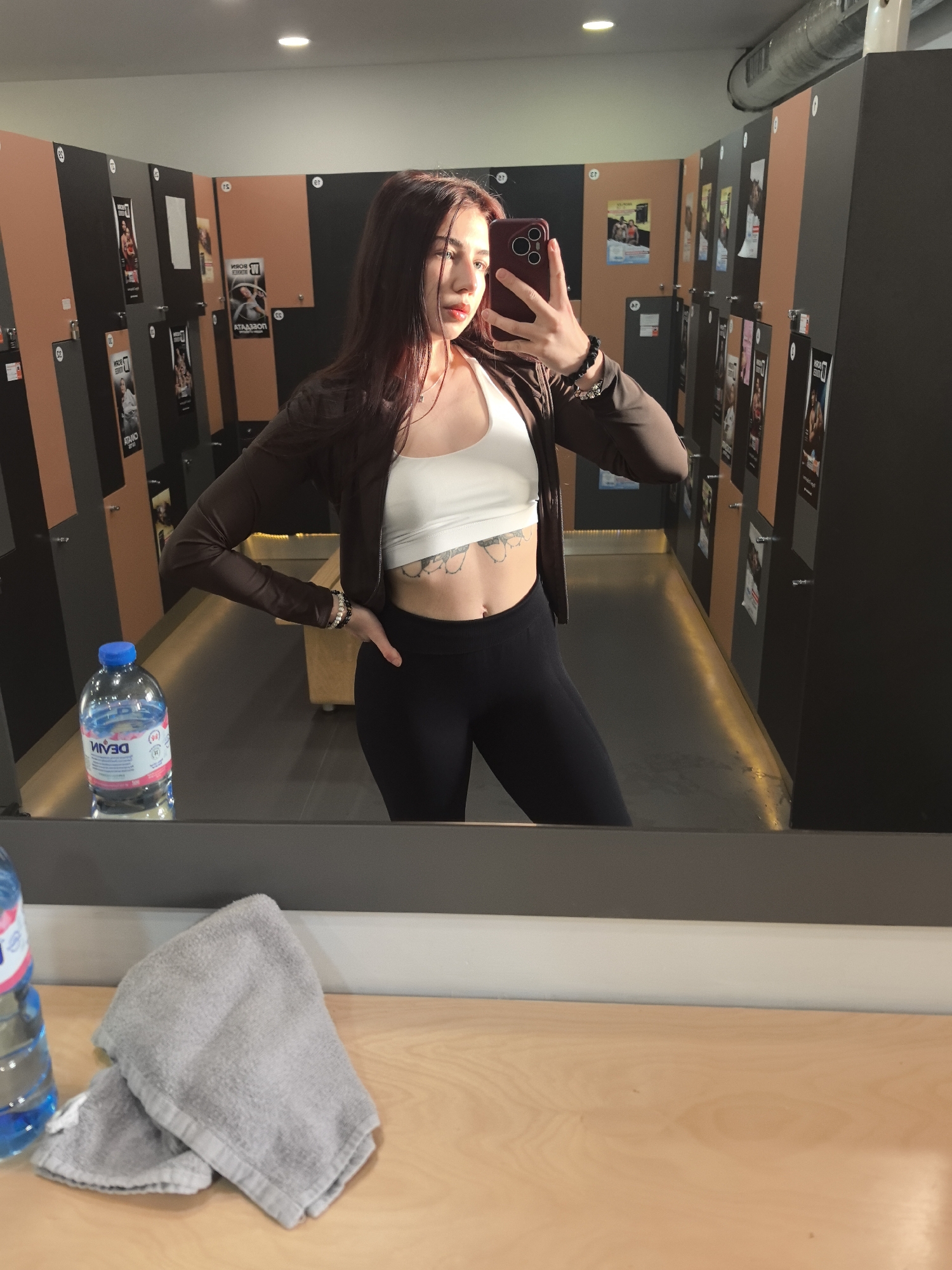 A woman is taking a selfie in a gym locker room. She is wearing a white sports bra, black leggings, and a brown jacket. She has red lipstick and dark hair. She is holding a red phone in her right hand. A water bottle and a folded gray towel sit on a wooden counter in front of the mirror. Behind her are lockers with advertisements posted on them. The gym is likely located in Stara Zagora, Bulgaria.
