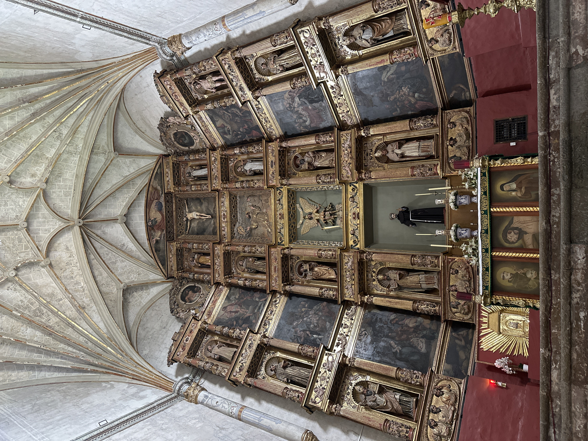 The image shows the interior of a church in Huejotzingo, Mexico, specifically focusing on a large, ornate altarpiece. The altarpiece is highly decorated with gilded wood, featuring numerous niches holding statues of saints and religious figures. Painted panels depicting biblical scenes are integrated into the structure. In the center of the altarpiece, a figure of Christ on the cross is prominently displayed. Below this, a statue of a robed male figure stands in a niche. The ceiling of the church is visible at the top, displaying intricate ribbed vaulting with gilded detailing. In the foreground, the altar itself is visible, adorned with paintings and religious artifacts, including candles and floral arrangements. The walls of the church are painted a deep red. The overall scene conveys a sense of historical and religious significance.