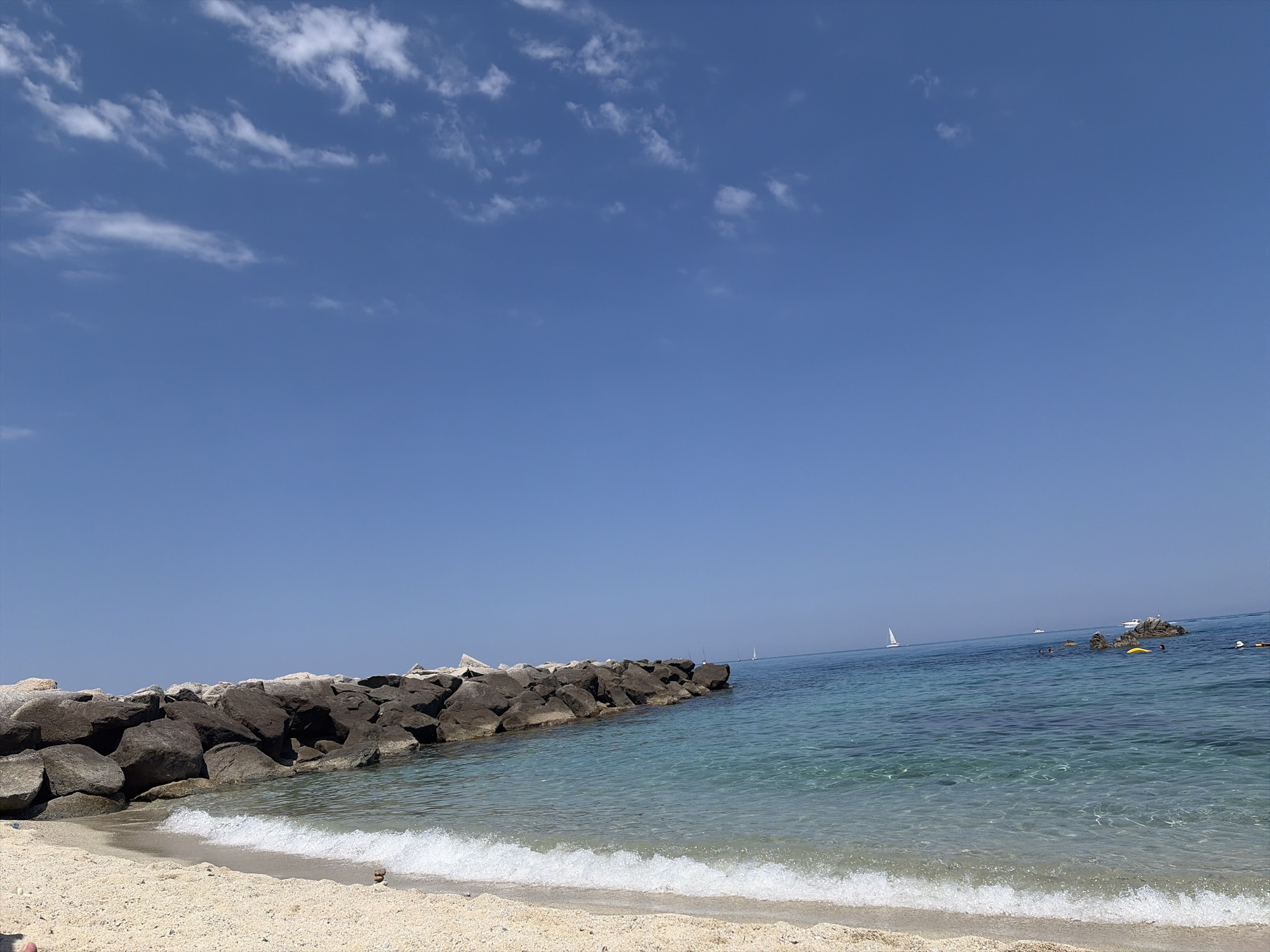 The image depicts a sunny day at the beach in Parghelia, Italy. A breakwater made of large, dark rocks extends into the clear, turquoise water. The sand is light-colored and textured, with a gentle wave breaking on the shore. In the distance, several sailboats can be seen on the horizon, and a few people are visible in the water. The sky is a vibrant blue with scattered white clouds.
