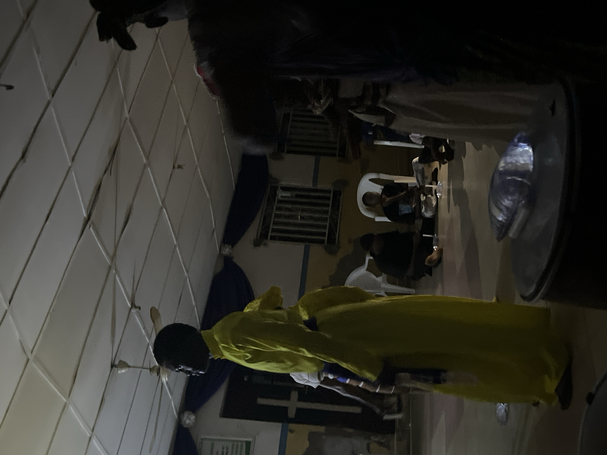 The image depicts an indoor gathering in a dimly lit space, with prominent shadows. In the left foreground, a person with dark skin and a clean-shaven head stands in profile, facing right. This individual wears a long, loose-fitting, bright yellow robe, seemingly cinched at the waist with a blue band. A small, reflective object, possibly a plastic-wrapped item, rests on the floor near their feet.
Behind the yellow-robed figure, in the left mid-ground, another person in a white top and dark lower garment is partially visible. In the extreme right foreground, a largely obscured figure, mostly in deep shadow, shows a dark, voluminous hairstyle and portions of a dark garment.
In the central and right mid-ground and background, several individuals are present. One person is seated on a white plastic chair to the right of the yellow-robed figure, wearing a dark top. Other figures are seated or kneeling on the light-colored floor, some partially obscured by chairs or shadows. White plastic chairs are scattered throughout the space.
The walls are painted with a tan or light yellow lower section and a light blue stripe or upper section, topped by dark blue fabric drapes along the upper perimeter. A green and white sign is partially visible on the far left wall. A distinctive white cross is painted on a dark door or wall panel in the left background. Barred windows are visible on the back wall.
The ceiling consists of white grid-style tiles, from which two multi-bladed ceiling fans are suspended. The floor is a light-colored, possibly tiled or concrete, surface. Artificial light sources from above illuminate the room, casting strong shadows, particularly on the right side and on the faces of individuals. The overall scene suggests a community or religious gathering in the city of Unknown, Unknown.