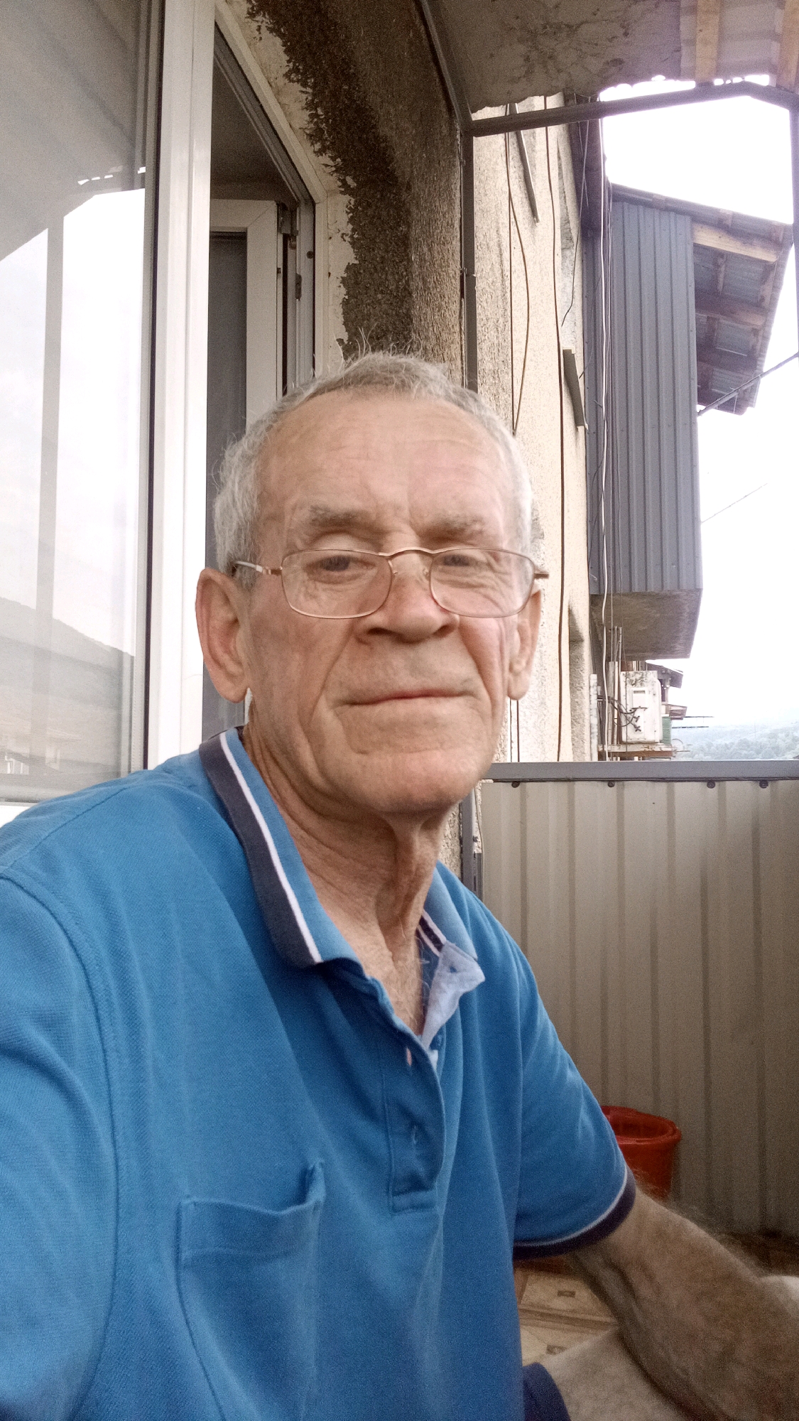 An elderly Caucasian man with graying hair and glasses is pictured in a close-up shot, looking directly at the camera. He is wearing a blue polo shirt with a white and dark blue striped collar. The man is outdoors, on a balcony or patio, with a building and some wires visible in the background. The scene suggests a casual, everyday moment. The overall lighting is soft, and the background is slightly blurred, drawing attention to the man's face. The image appears to be a selfie taken by the man himself. The backdrop suggests an urban or semi-urban environment, possibly a residential area in Tiachiv, Ukraine, given the context.