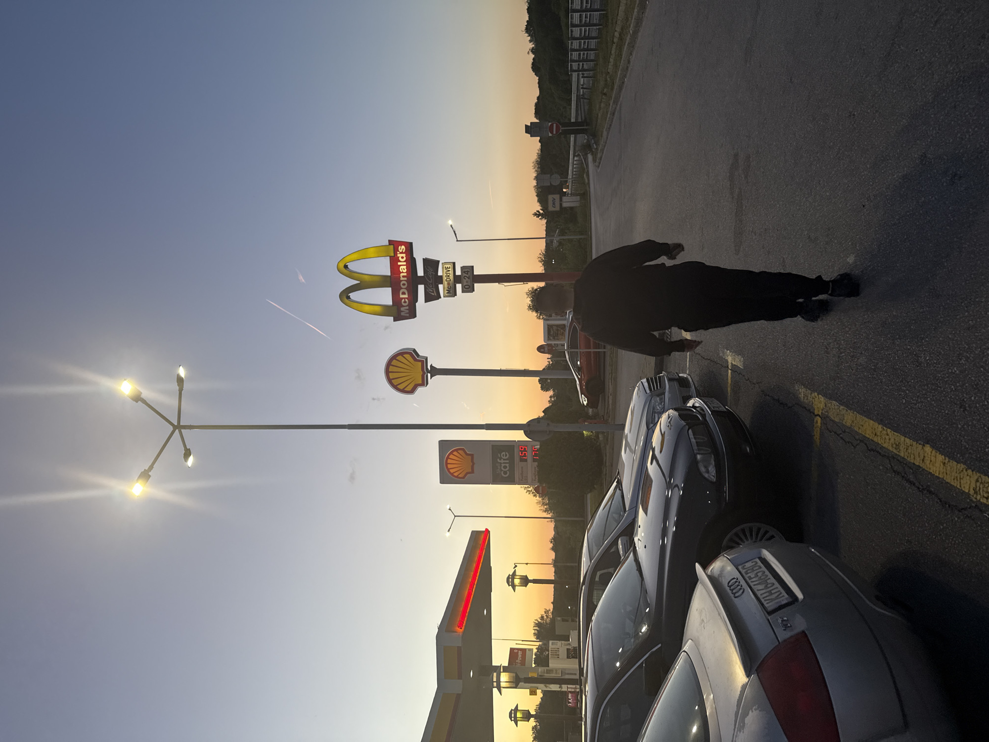 The image shows a roadside scene in Studena, Bulgaria, at dusk or dawn. A person in dark clothing is walking away from the camera on an asphalt surface marked with yellow lines, near parked cars. In the background, there are McDonald's and Shell signs, with gas prices visible as 1.59. Further back, trees are visible against the horizon, beneath a sky with an orange to blue gradient. A light post with multiple lamps is prominently positioned near the McDonald's sign.
