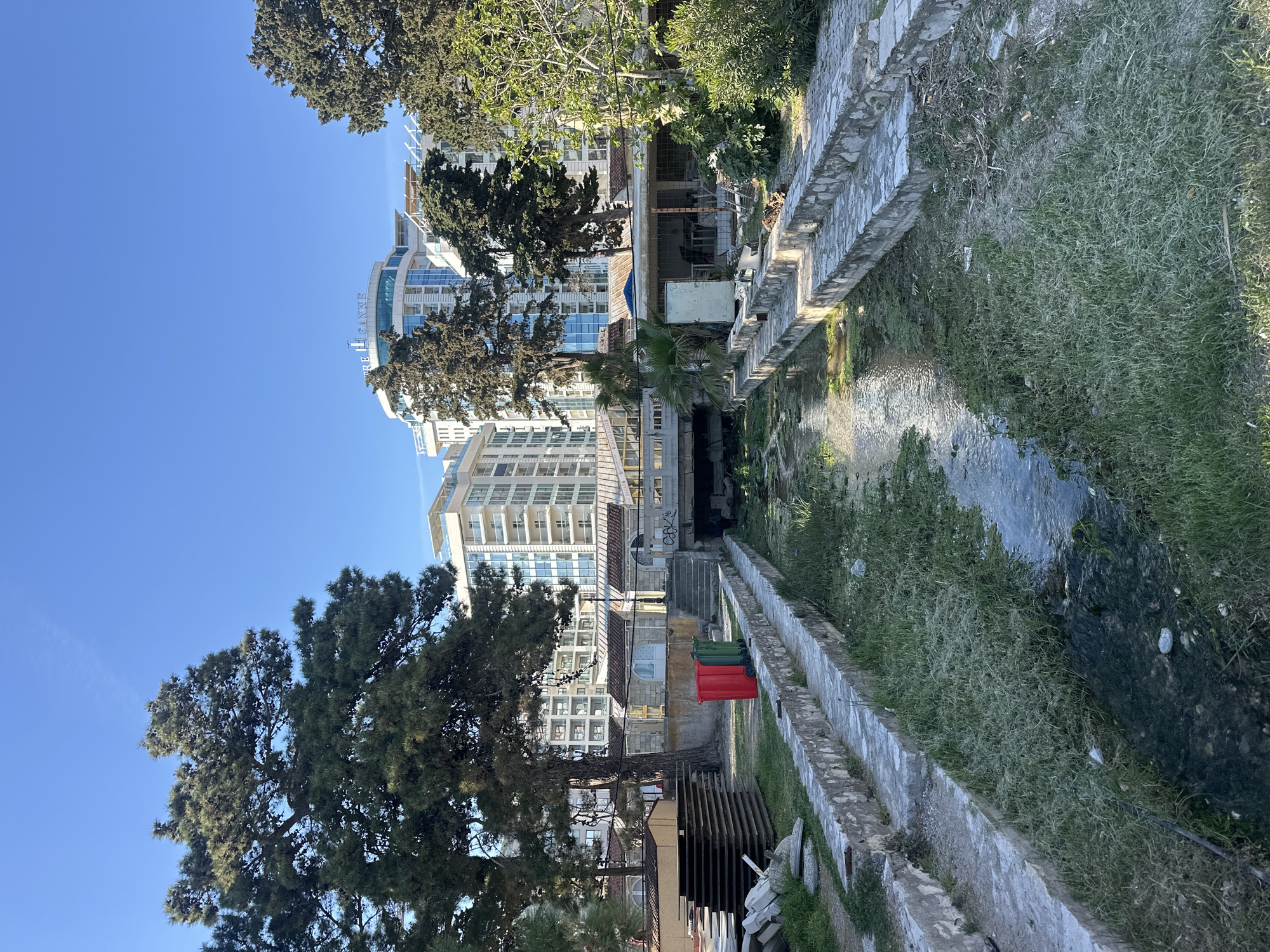 The image depicts a small, concrete-lined stream or drainage canal in what appears to be an urban setting. The canal is bordered by grassy areas on either side, and a stone wall runs along its edge. In the background, several multi-story buildings are visible, including a high-rise hotel or apartment complex with a glass facade. The sky is clear and blue. There are some trash containers. The scene seems to be located in the city of Budva, Montenegro.